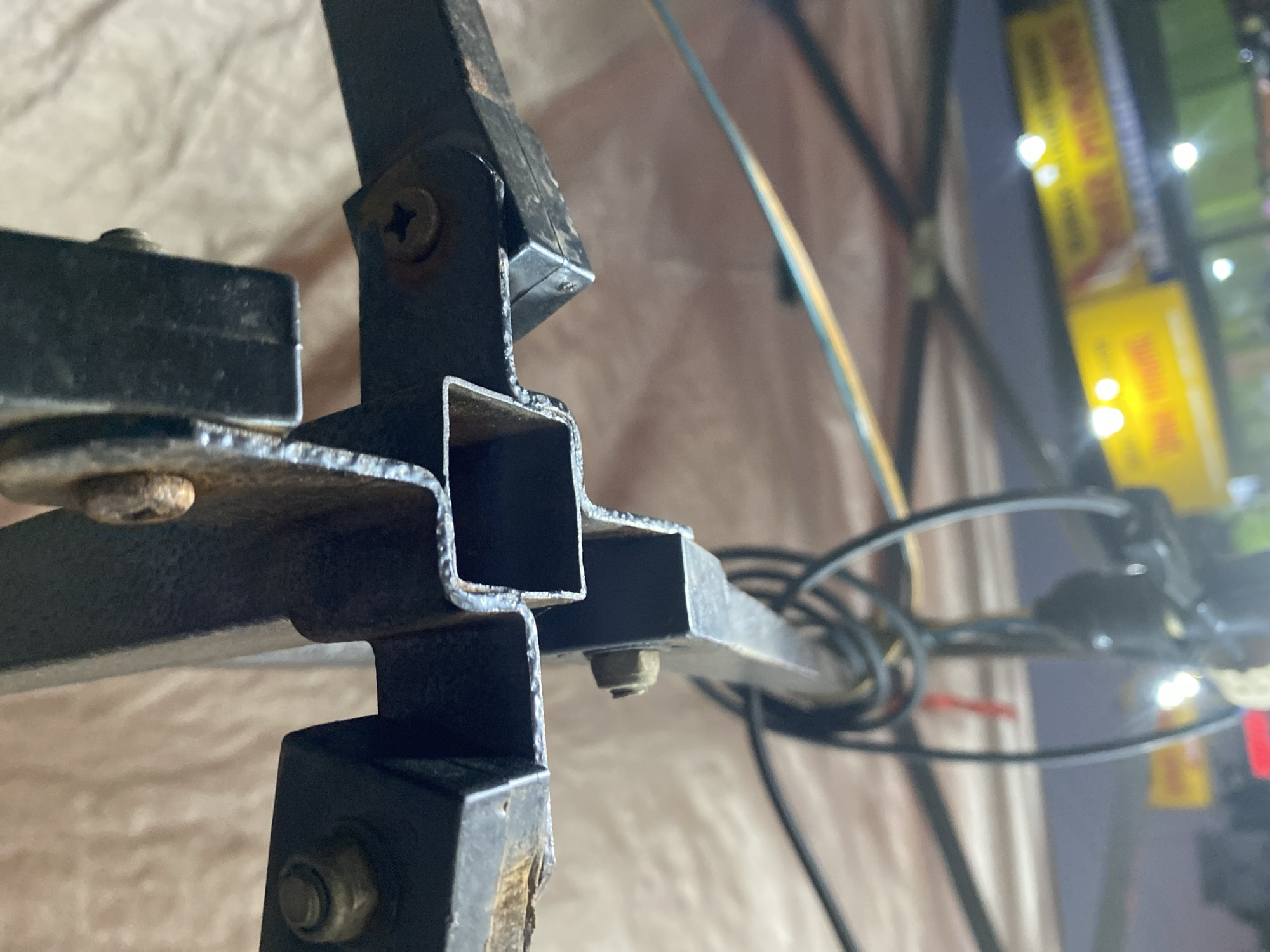 This is a close-up, low-angle, eye-level shot of the underside of a metal structure, likely part of a tent or canopy, in Kasihan, Indonesia. The focus is on the metal frame, which appears to be made of black painted square tubing joined by bolts and screws. There are signs of rust and wear on the metal. A light-colored fabric, possibly the canopy material, is visible in the background. Some black electrical cables are coiled and hanging beneath the structure. In the background, out of focus, are bright yellow signs with text in an Indonesian script, suggesting a commercial or market area, possibly illuminated by lights. The overall impression is of a functional, outdoor structure in a somewhat weathered condition.