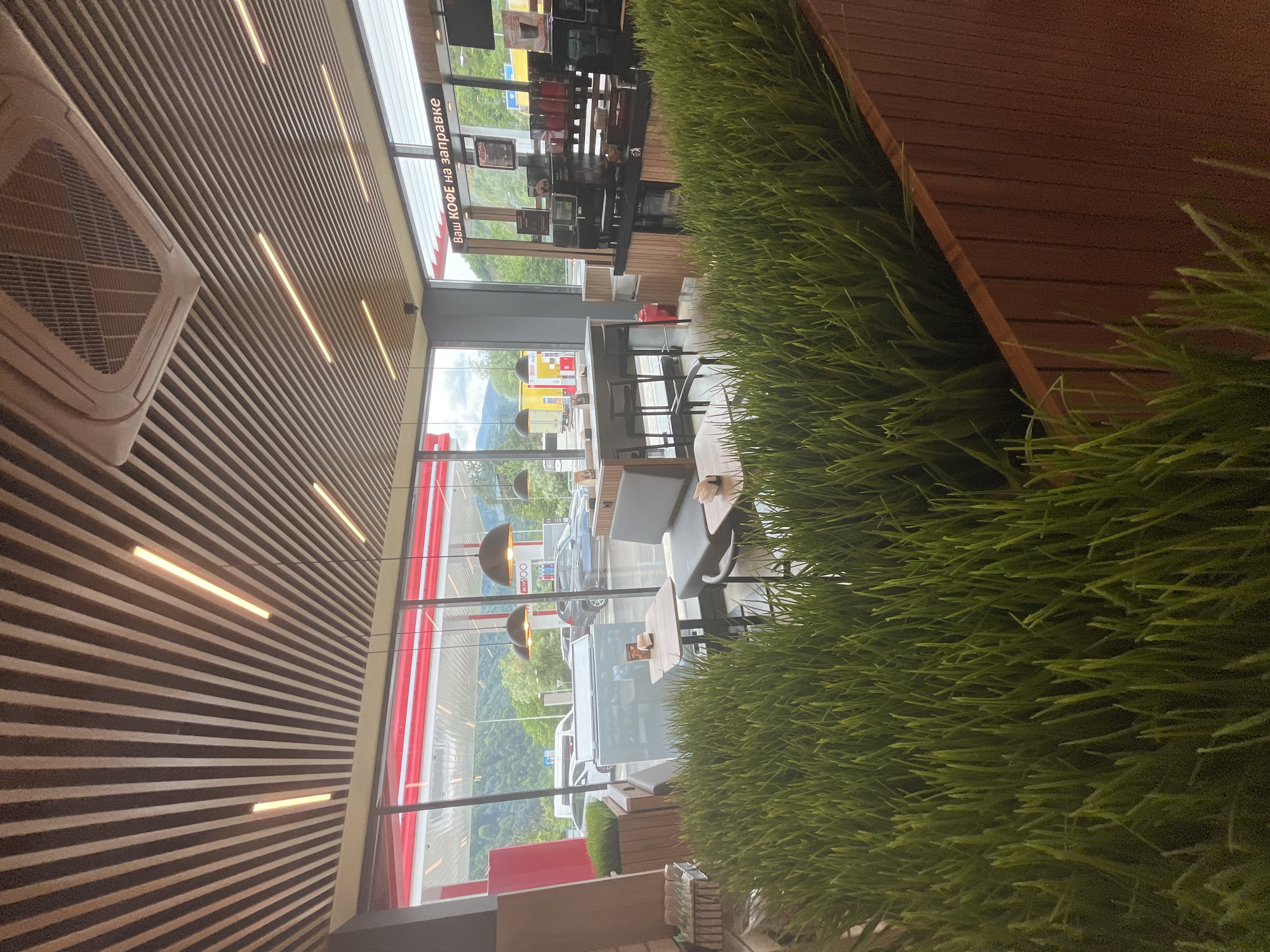 The image depicts the interior of a modern cafe situated at a gas station in Krasnaya Polyana, Russia. In the foreground, a large planter filled with vibrant green artificial grass creates a naturalistic divider. Beyond the grass, several cafe tables with chairs are visible, some with patrons. Through a large window, a gas station canopy with pumps and cars can be seen, along with a mountainous, wooded landscape in the background under an overcast sky. The cafe interior features a contemporary design with a slatted wooden ceiling, recessed lighting, and a coffee preparation counter. A sign above the counter reads "Ваш КОФЕ на заправке," which translates to "Your Coffee at the Gas Station."