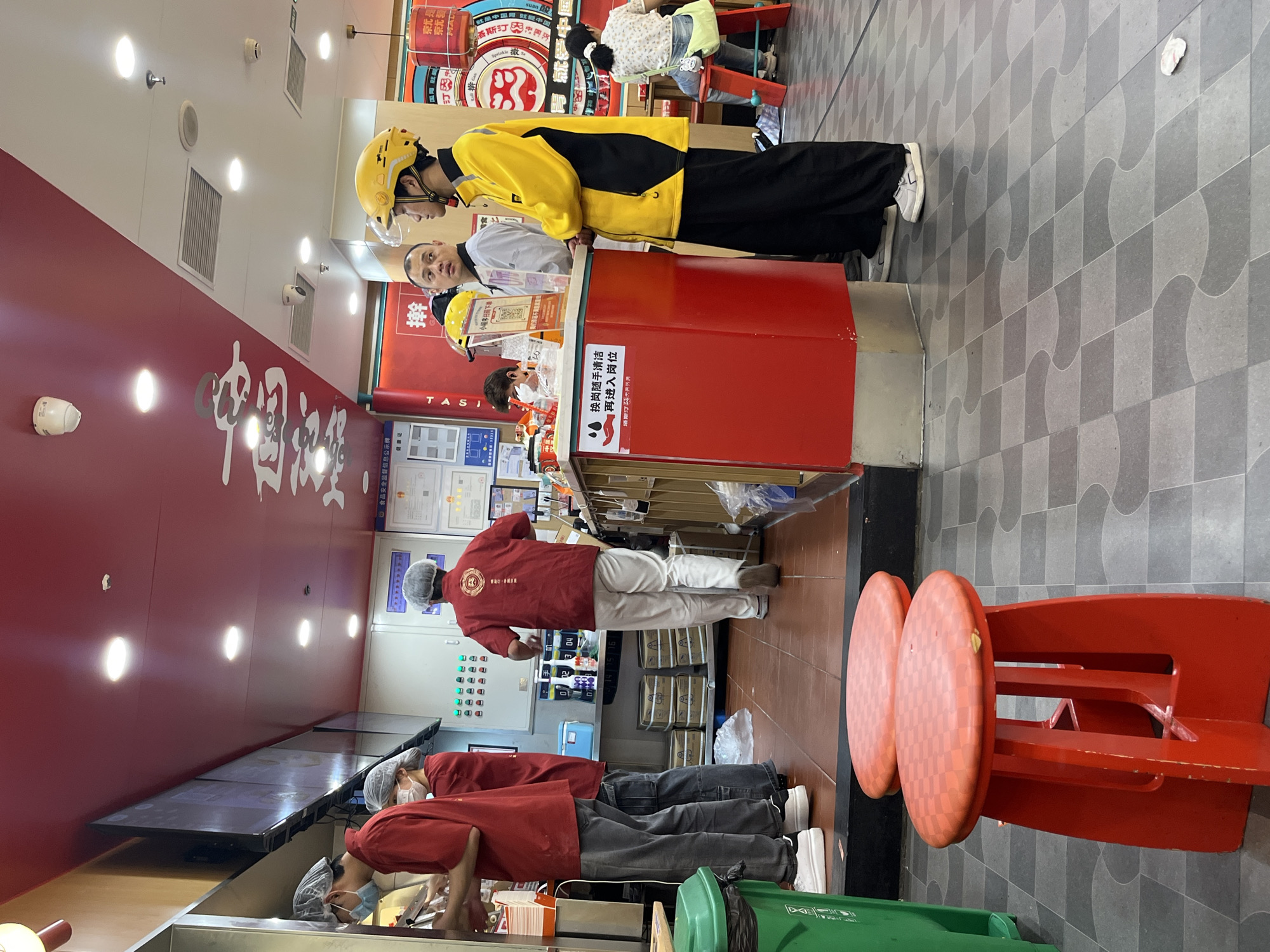 This is an indoor scene, likely a food establishment in Yuzhong District, China, characterized by vibrant red accents and tiled flooring. In the foreground, two bright red stools with checkered tops are positioned on a grey and dark grey patterned tiled floor. To the left, a tall green trash receptacle stands near the entrance.
The mid-ground reveals a service counter area. Several individuals are present. Two people in red uniforms and hairnets are attending to customers or preparing orders behind the counter. One appears to be holding a utensil and interacting with something on the counter, while the other is facing away from the camera. Further to the right, two individuals in yellow delivery uniforms, one wearing a yellow helmet, are standing at the counter, possibly waiting for orders or interacting with staff. A man in a white shirt is also visible behind the counter, speaking with the delivery personnel.
Behind the counter, stacked boxes and shelves with various items, including what appear to be bottles and containers, are visible. A sign with Chinese characters and a QR code is mounted on a red surface.
In the background, more of the establishment's interior is visible, including a red ceiling with recessed lighting and some decorative elements with Chinese calligraphy. A woman is seated on a red stool in the far right, partially visible. The overall impression is a busy service environment.
