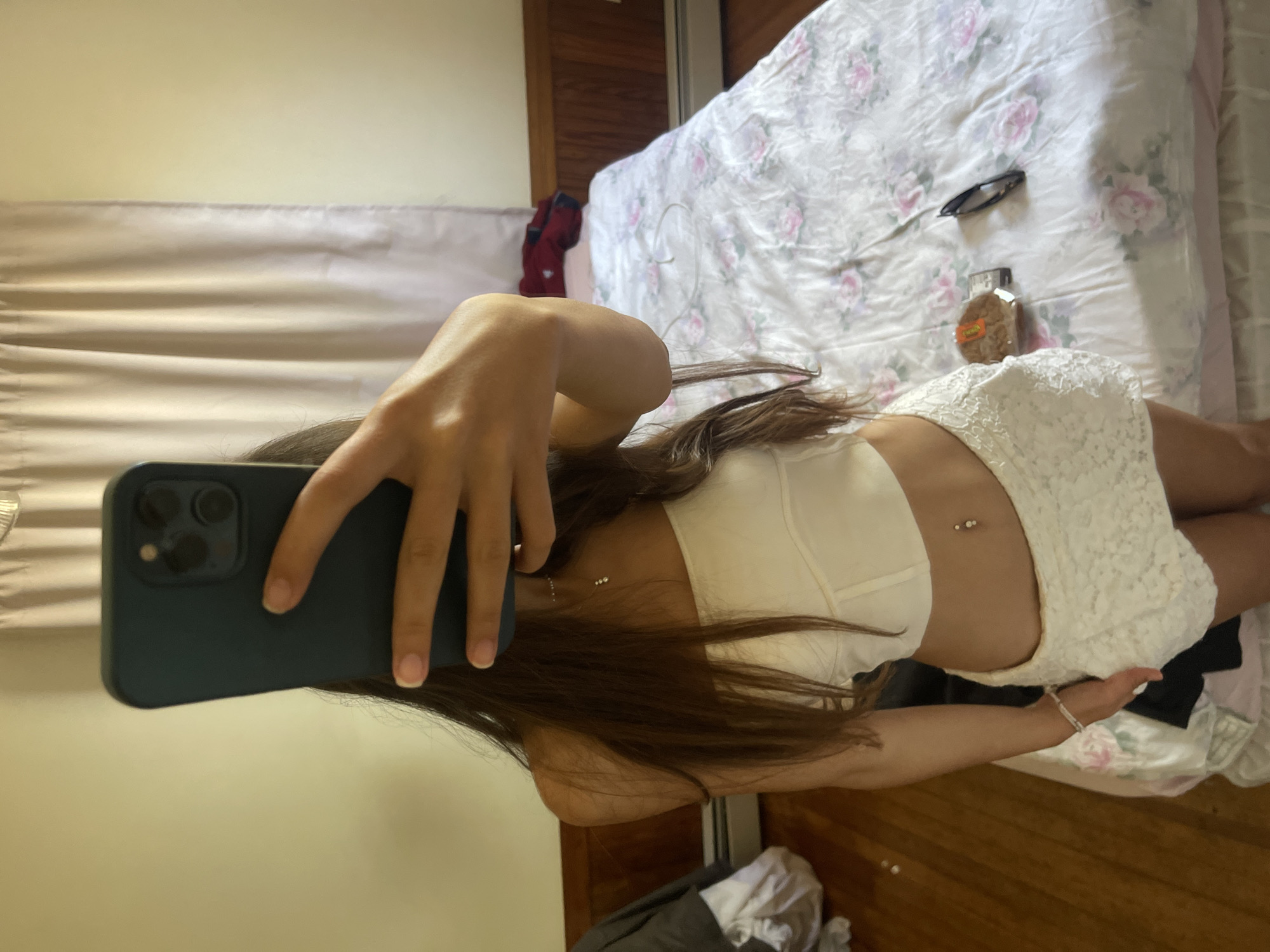 A young woman is taking a mirror selfie in a bedroom. She is wearing a white tube top and a white lace skirt. She has long brown hair and a belly button piercing. The room has wood floors, a bed with floral bedding, and a radiator. There are curtains on the wall and a dresser with clothing on it. The image was taken in Oak Bluffs, United States of America.