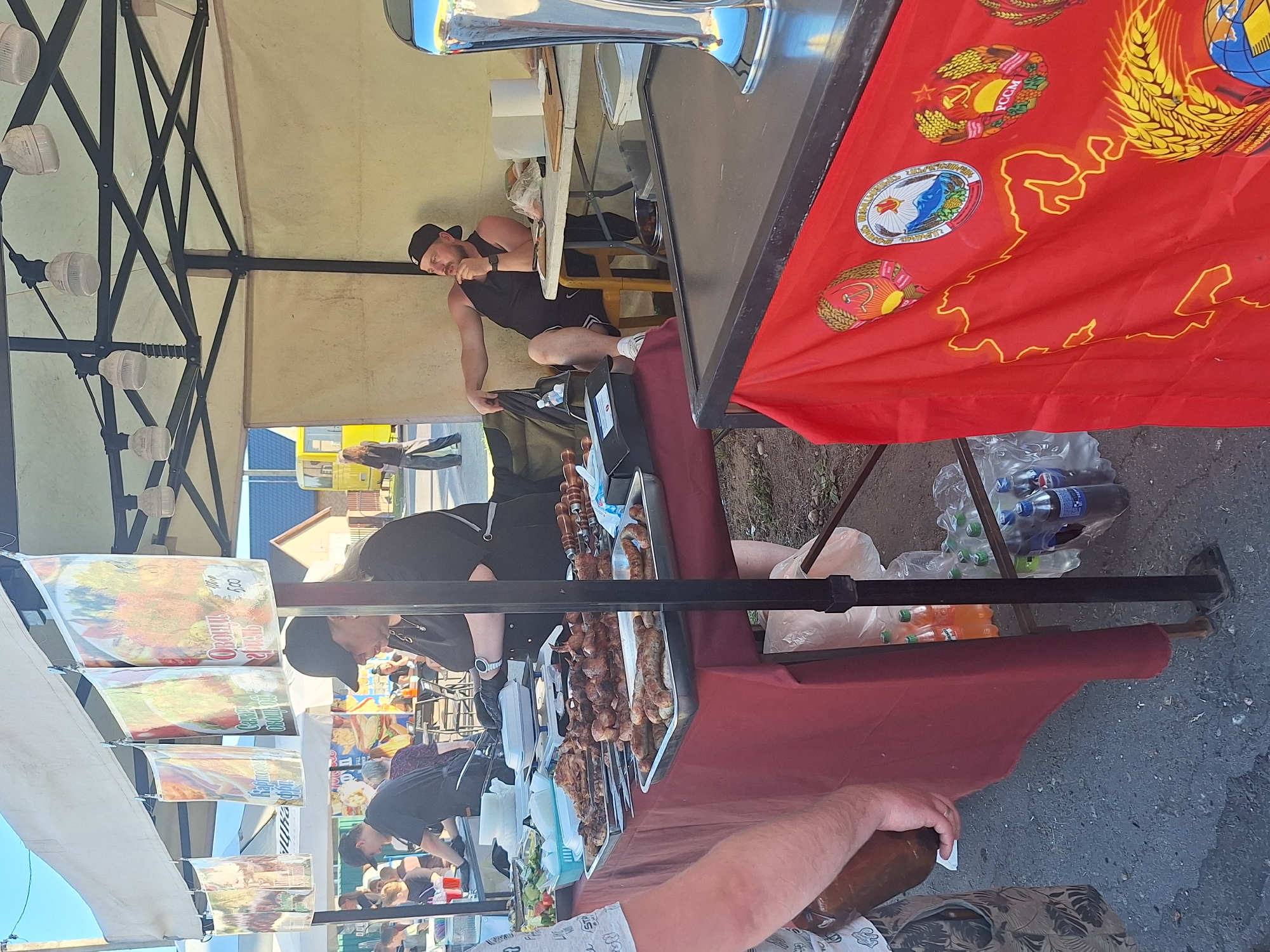 At an outdoor market in Цімкавіцкі сельскі Савет, Belarus, a food stall displays trays of grilled meats and sausages. Two vendors, a woman in a black cap and apron and a man wearing a black tank top and cap, are present. The woman is behind the counter, seemingly preparing food, while the man sits in a chair, looking thoughtfully to the side. Various beverages are visible in plastic bags and on shelves. A red banner with Soviet-era emblems and a map of the USSR hangs from the front of the stall. The background shows other market stalls, people, and a yellow bus. The overall scene depicts a bustling outdoor food vendor operation.