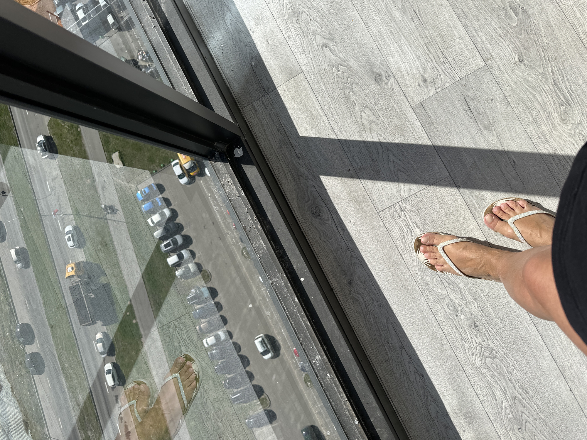 The image depicts a high-angle view from a window, possibly in a high-rise building, in the city of Unknown, Russia. The viewpoint shows a reflection of the person's feet wearing white flip-flops on the windowpane, alongside the view of the street below. Outside, there is a street with multiple lanes, and a row of parked vehicles. There is green space adjacent to the street. Inside, the person is standing on light-colored hardwood flooring. A shadow cast by the window frame bisects the flooring diagonally.