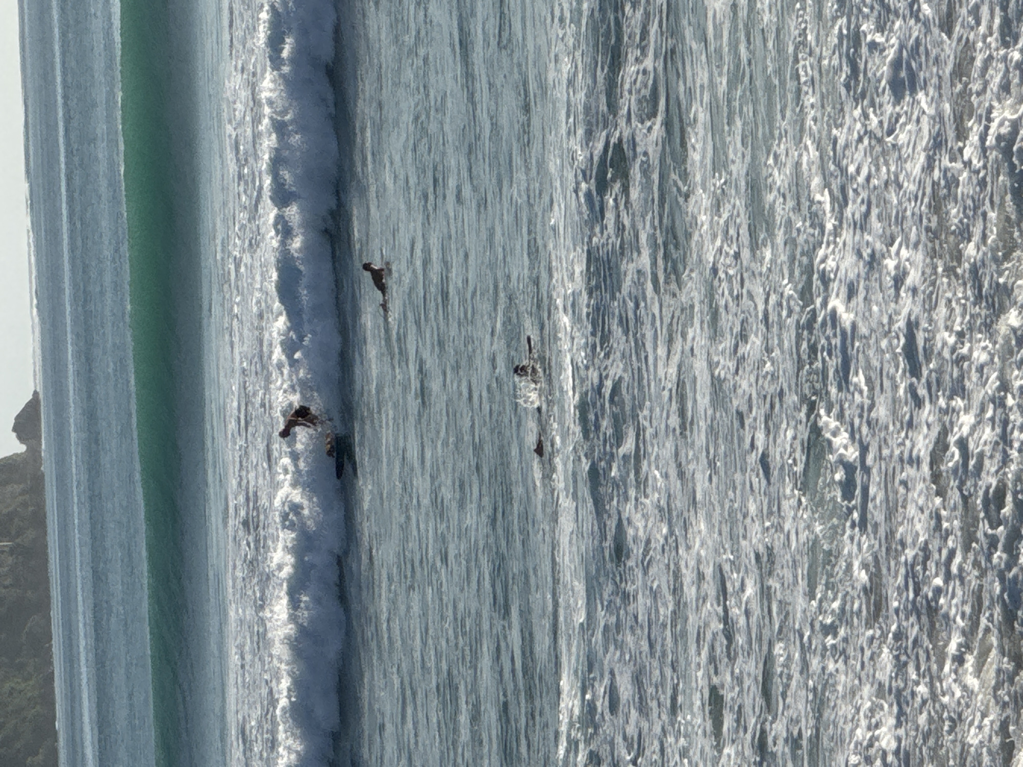 The image depicts an aerial view of the ocean in Kuta Selatan, Indonesia. Several surfers are visible, riding waves near the shore. The water is turbulent with a mix of green and blue hues, exhibiting white foamy waves. Along the left edge of the frame, land is visible, displaying a rocky outcrop and vegetation.
