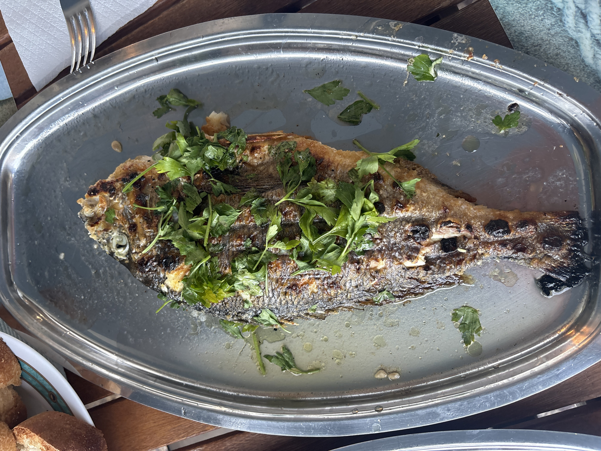A whole, grilled fish is presented on a silver platter, garnished with fresh parsley. The fish appears to be a whole sea bream, grilled to a golden-brown perfection, with visible grill marks. Its scales are intact, and its eye is clearly visible. The platter is oval and made of polished metal, with a slightly raised rim. Scattered around the fish are droplets of oil and small pieces of parsley. To the upper left of the platter, a white plate holds several slices of toasted bread. The entire scene is set on a wooden table, suggesting an outdoor dining setting, possibly in Mandra, Greece. A fork is visible on the right side of the image, resting on a white napkin. The lighting suggests it is daytime. There are no people visible in the image.