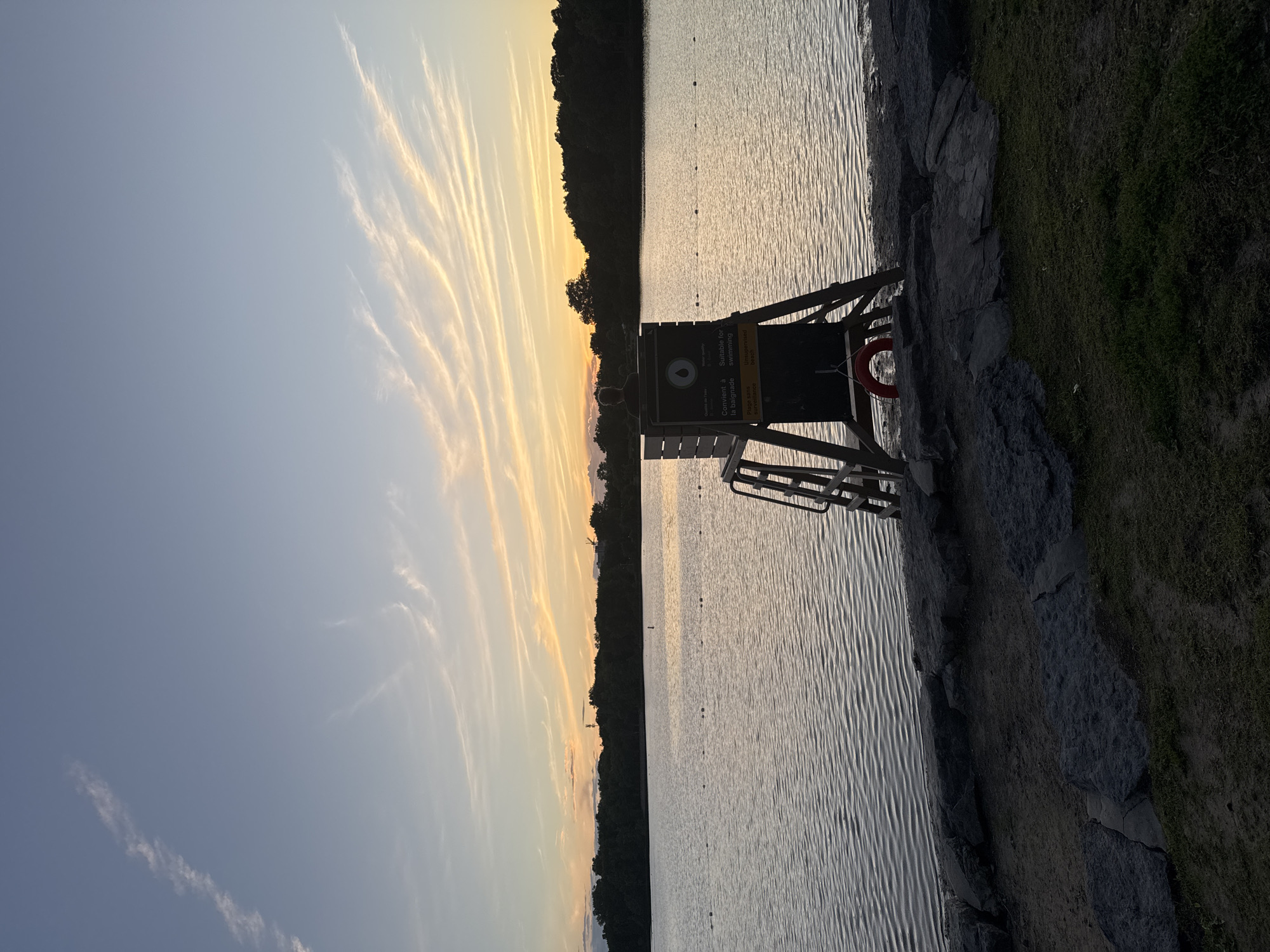 A lifeguard chair stands on the shore of a calm lake in Gatineau, Canada, at sunset. The sky is a gradient of pale blue to soft orange and yellow, with wispy clouds streaked across it. The water reflects the sky and is bordered by a line of dark trees in the distance. The lifeguard chair is wooden with a white box at the top, where a person is silhouetted against the sky. A red life preserver hangs from the side of the chair. The foreground consists of rocks and grass.