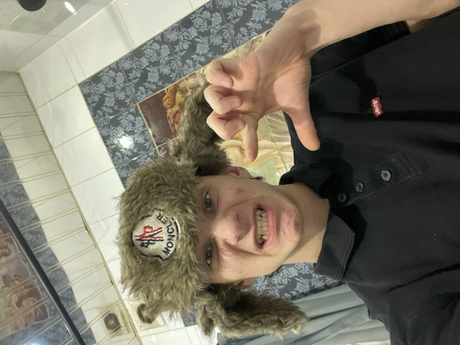 A young man poses for a selfie, wearing a Moncler furry trapper hat and a black polo shirt with a red Levi's tag on the chest. He is in what appears to be a tiled bathroom, as indicated by the patterned tile wall behind him. He is grimacing and making a claw gesture with his right hand. The photo is taken at an unusual angle, with the camera tilted to the left.