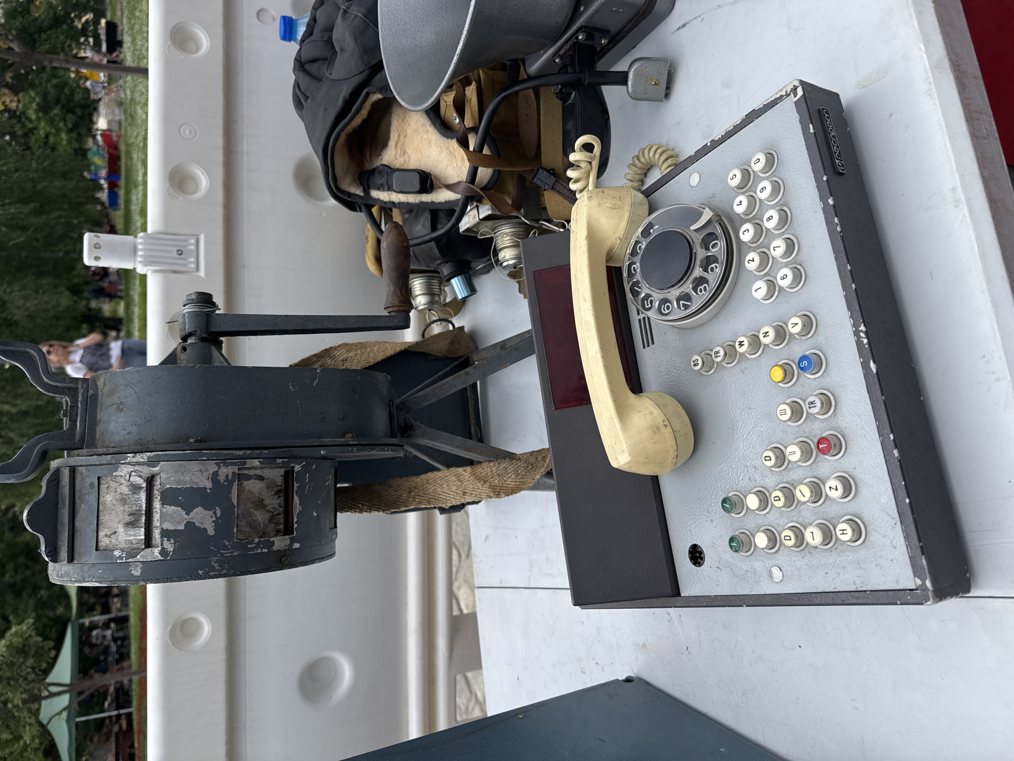 The image shows a collection of vintage military equipment, including a rotary and push-button telephone switchboard with Cyrillic labeling, possibly Russian. The switchboard is gray with numerous white buttons and a yellow handset. Next to it is a headgear, possibly a pilot's helmet or headset, with fur lining and straps. There is also a metal apparatus that appears to be a rotating mechanism with an attached handle. The equipment is placed on a white surface with a park visible in the background. The location is unidentified.