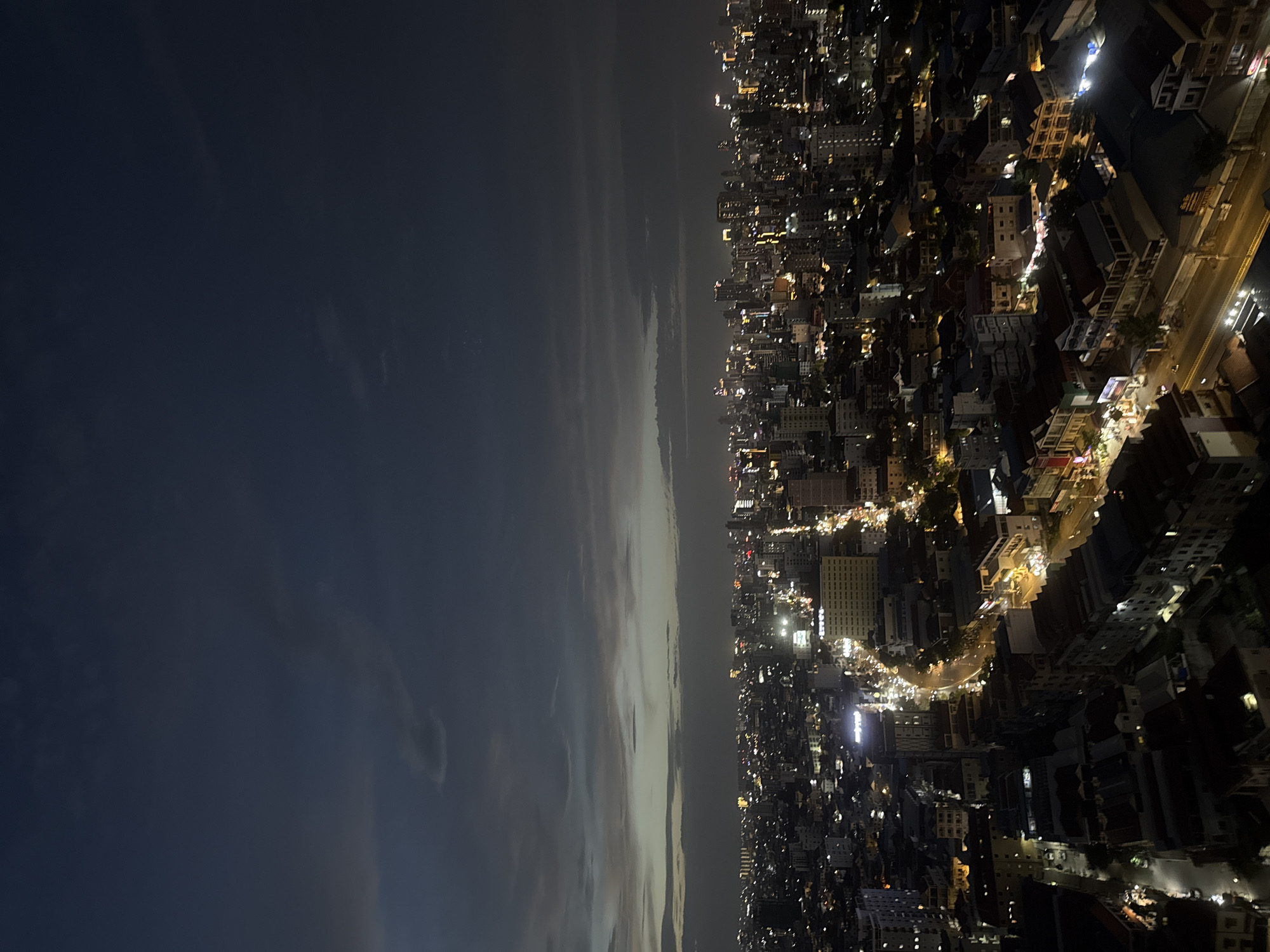 The image is an aerial nighttime view of a densely populated urban area, likely Khan Mean Chey, Cambodia. The scene is dominated by a sprawling cityscape illuminated by numerous artificial lights from buildings and streetlights. The sky above is dark with scattered clouds, suggesting it may be dusk or nighttime with some ambient light pollution. The foreground shows the rooftops of buildings, many with visible windows and structures. Further back, the cityscape extends towards the horizon, with a mix of high-rise buildings and lower-density structures. Roads are visible, marked by lines of headlights and taillights from moving vehicles, creating streaks of light. Some buildings have illuminated signage. The overall impression is of a bustling urban environment at night.