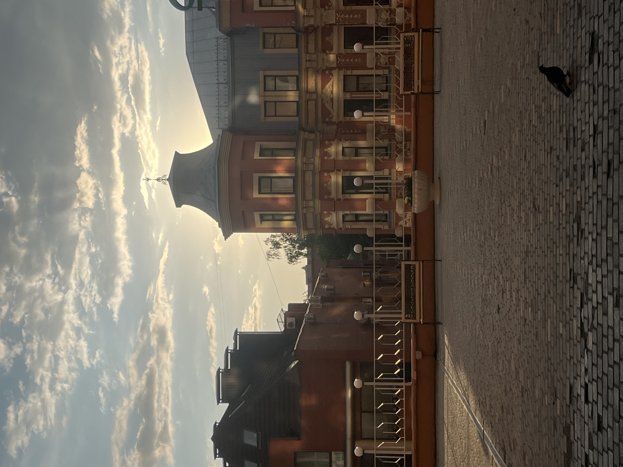 A vertical shot captures a brick-paved plaza and a multi-story building with ornate architectural details under a sky filled with scattered clouds during the golden hour. The building, painted in shades of red and adorned with decorative white trim, features numerous windows and balconies with intricate metal railings. Several round light fixtures are visible along the building's facade.
In the foreground, a single dark bird, possibly a pigeon, stands on the brickwork. The sky is a soft blue, transitioning to a warm yellow near the horizon, indicating either sunrise or sunset. A large, pale pot sits near the building's base, suggesting a decorative element. The overall scene is serene and architectural, with a touch of nature introduced by the bird and the visible trees peeking from behind the building. The image appears to be taken in Dnipro, Ukraine, based on the provided context.