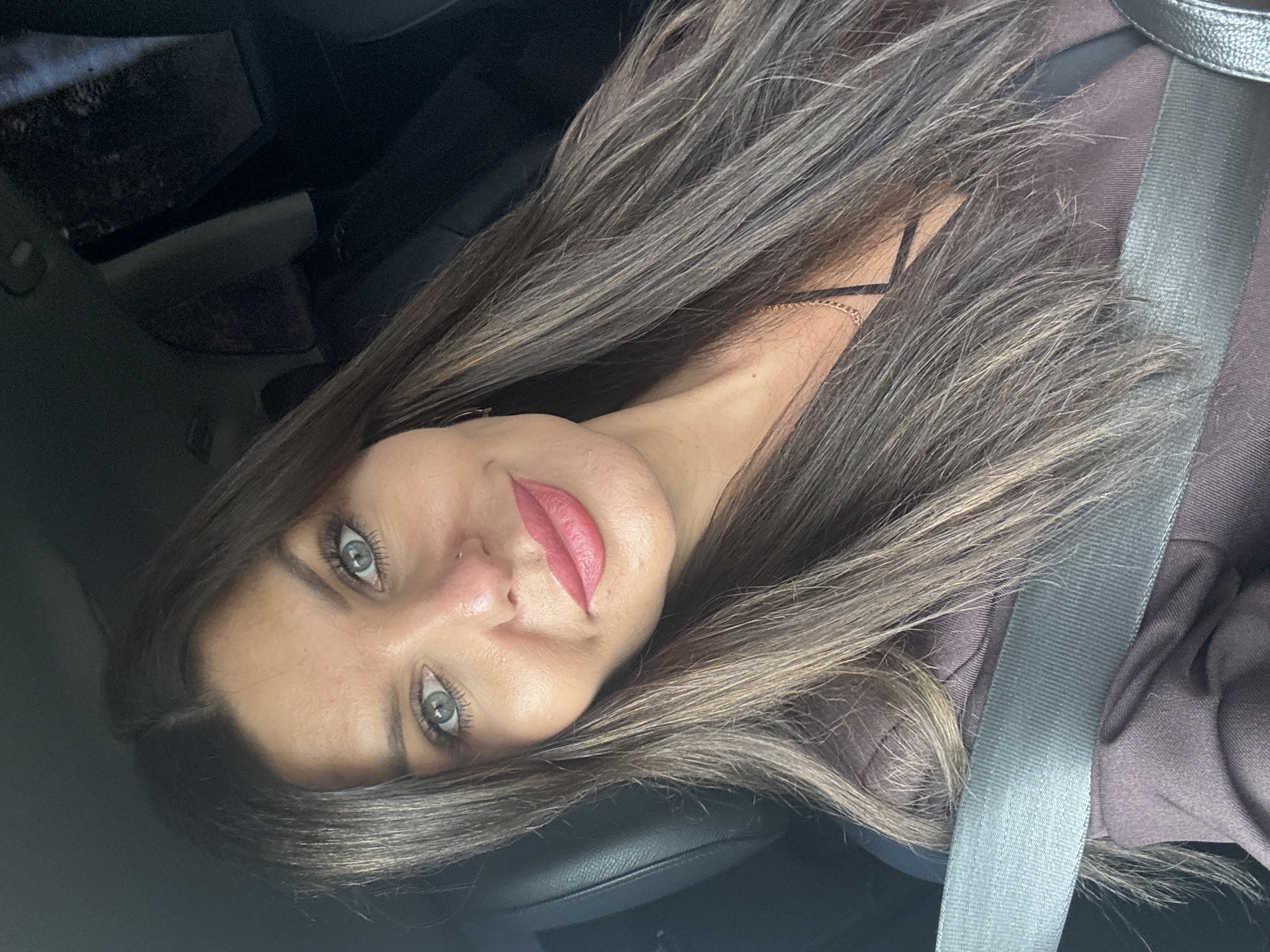 A medium shot of a woman wearing a brown top and a seatbelt, taken from a car. She has long, dark brown hair with lighter brown highlights, blue-green eyes, and pink lipstick. Her head is tilted, and she is looking at the camera. The background is dark, suggesting the interior of a car.