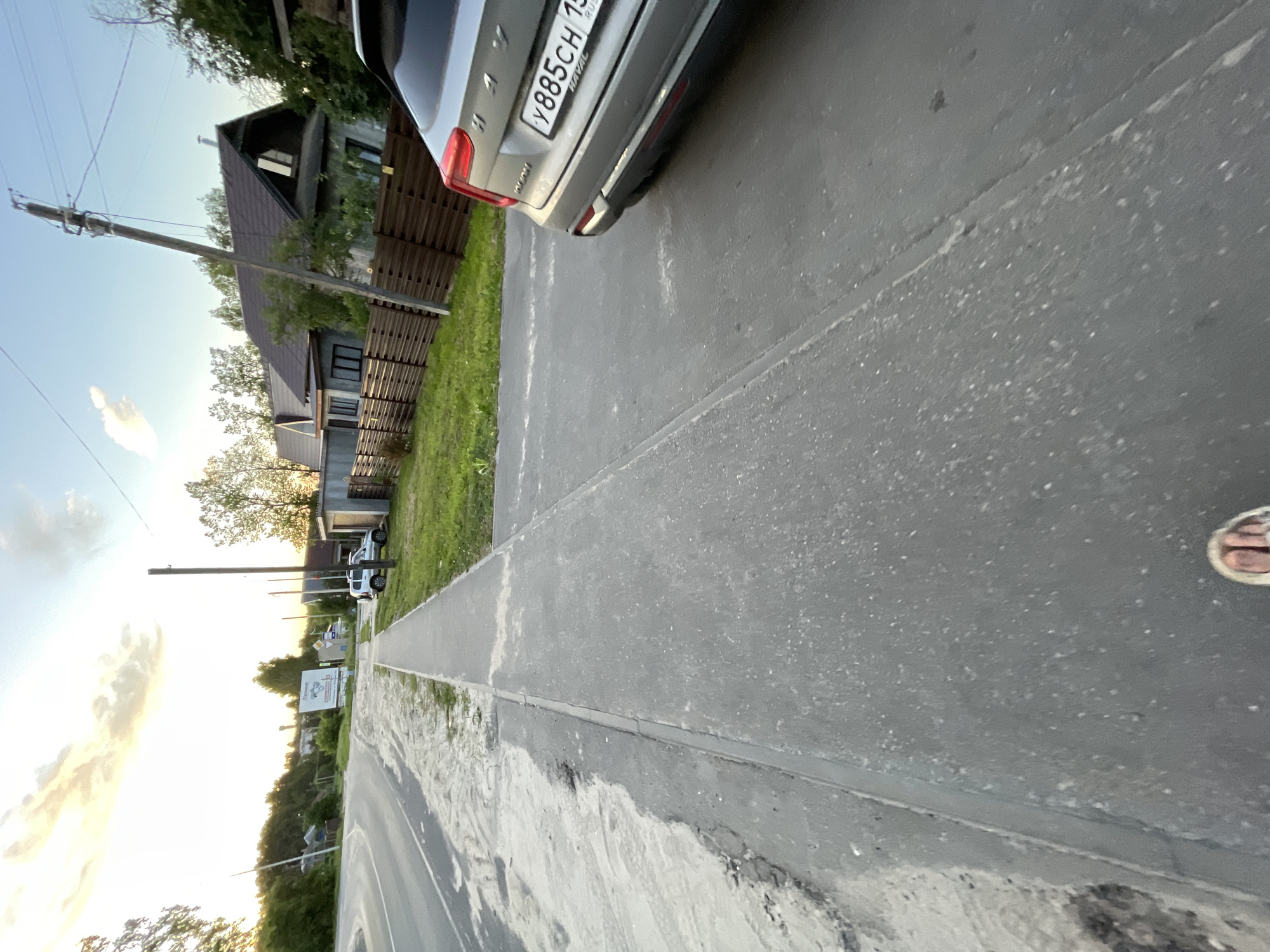 This image captures a street scene in Krasnye Baki, Russia. A grey Haval SUV with the license plate Y885CH 15 is parked on the right side of the road, partially obscuring a wooden fence. A sidewalk runs parallel to the road, with a portion of the road covered in sand and gravel. The background features several residential houses with dark tiled roofs, interspersed with trees. A power pole stands prominently in the middle ground. The sky above is clear with a few scattered clouds, and the sun appears to be setting, casting a warm glow. A person's foot in a sandal is visible in the foreground, suggesting the image was taken from a low vantage point, possibly while walking.