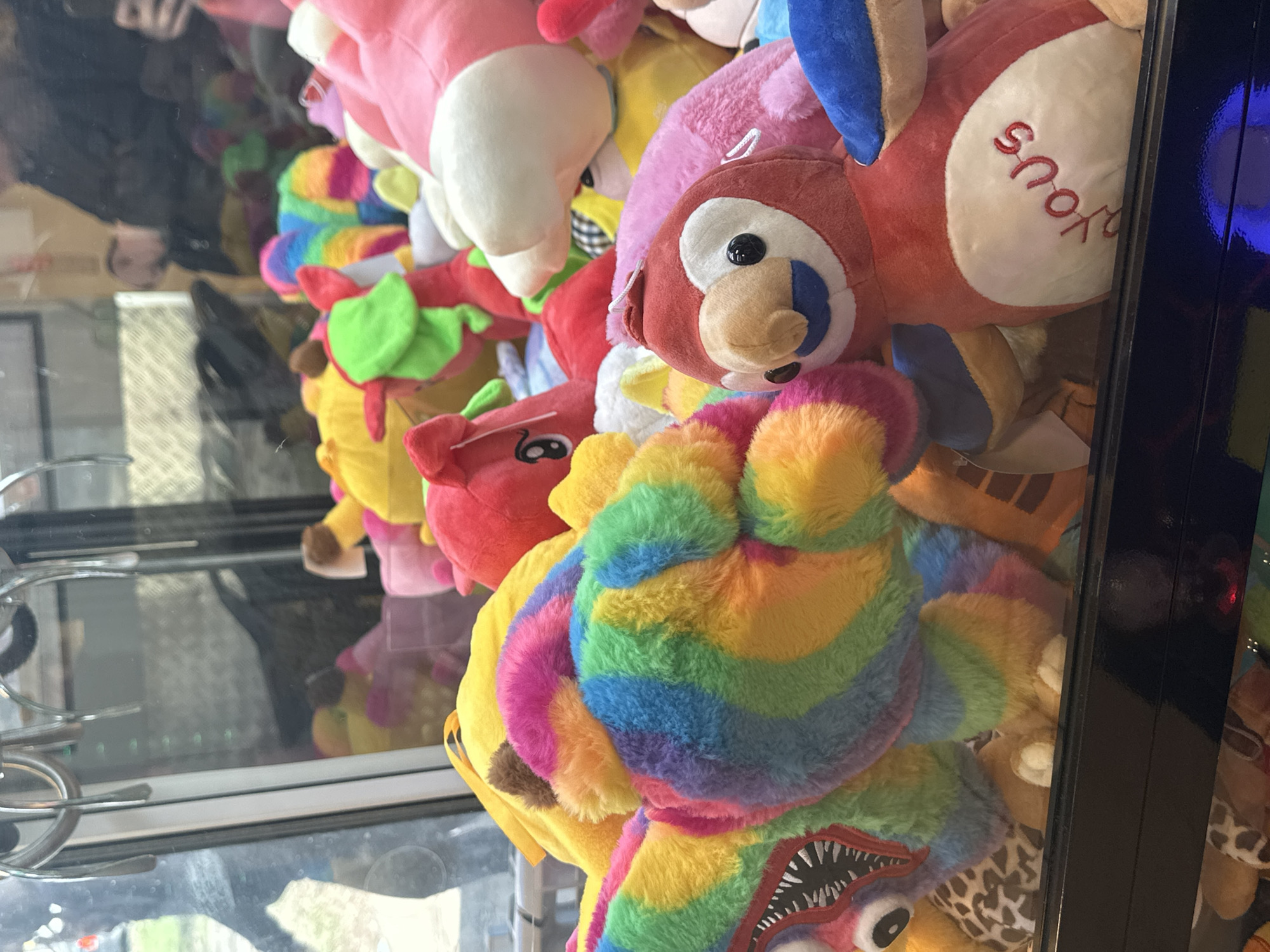 A claw machine, filled with stuffed animals, is captured in an eye-level, slightly skewed shot. The machine's metal claw arm is visible on the left side, with its reflection on the glass. The interior is packed with plush toys in various colors and shapes, including an owl with the word "you's" embroidered on its side, a pink pig, and a brightly colored rainbow-striped toy with large teeth and visible eyes. The machine itself is situated indoors, with reflections of the surrounding environment visible in the glass casing. The exact location in Moscow, Russia is not evident from the image.