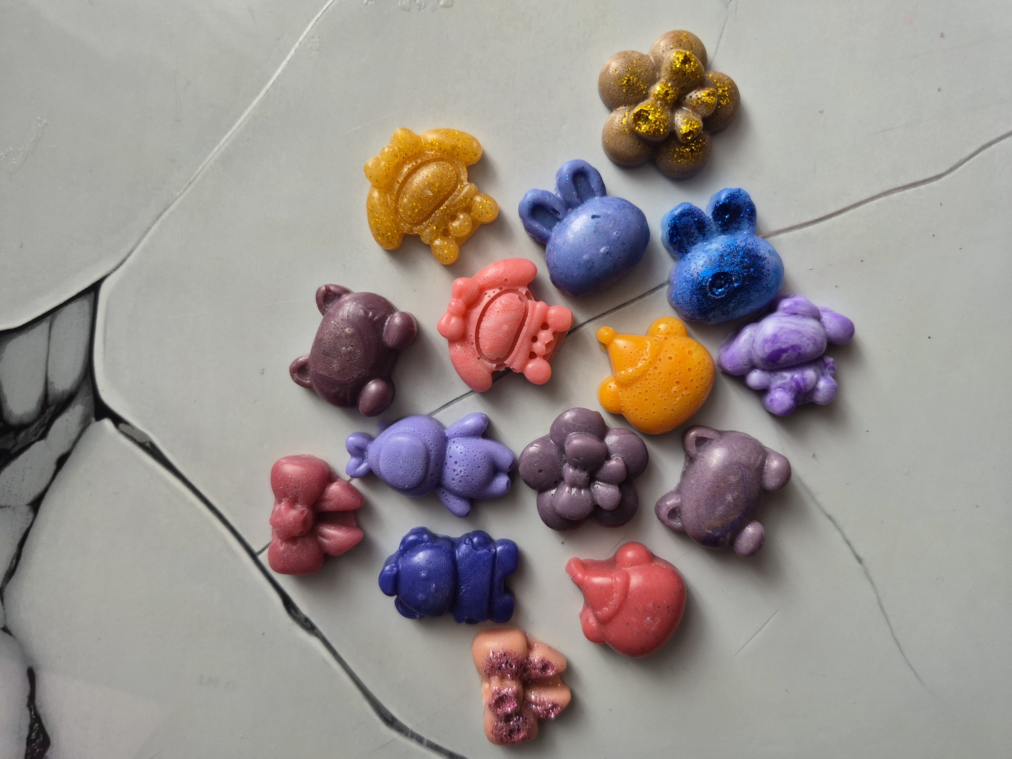 The image displays a collection of colorful, molded wax melts arranged on a light grey surface with subtle white marbling and black lines, possibly a countertop or tile. The wax melts are shaped into various forms, including teddy bears, bunnies, and flower-like clusters. The colors range from yellow, orange, pink, red, blue, purple, and brown, with some containing glitter or a shimmering effect. No people, actions, or interactions are visible in the image. The location cannot be determined from the provided image and has no bearing on its contents.
