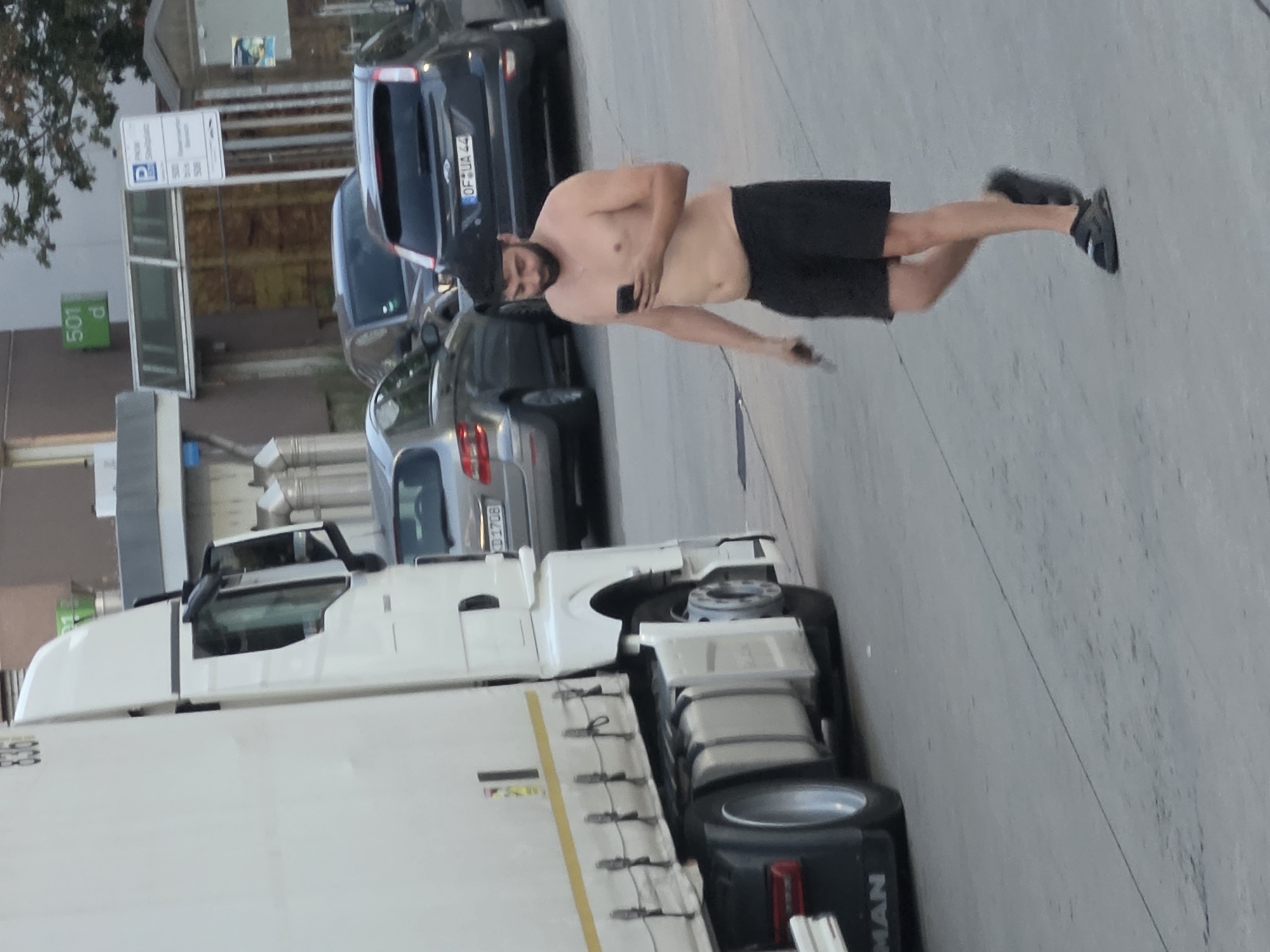 A topless man with a beard wearing black shorts and sandals walks across a paved area, looking at his phone. He is holding a small object in his left hand. Behind him, a white semi-trailer truck is parked. In the background, several cars are visible, including a dark grey Ford with a German license plate that reads "OF-UA 44". A parking sign with German text and a blue "P" symbol is also present, indicating a parking area in Frankfurt, Germany. The environment appears to be an urban setting with buildings and trees.