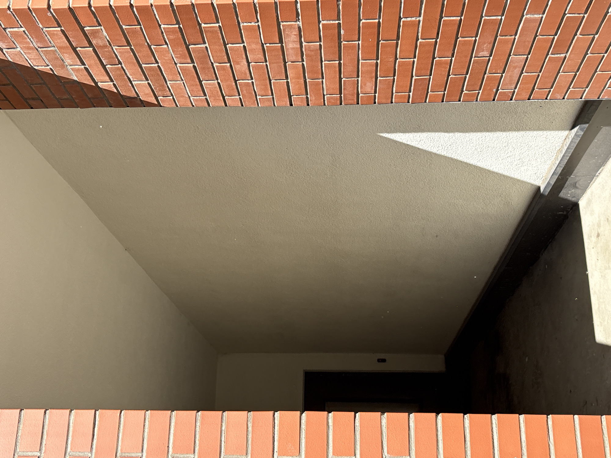 An exterior view of a building entrance, possibly a residential building, with red brick pillars on either side of a hallway. The hallway has light-colored walls and a dark, possibly concrete floor. Sunlight casts a sharp shadow on the wall from the right brick pillar. A dark doorway is visible in the background on the left side of the hallway. The scene is devoid of people.