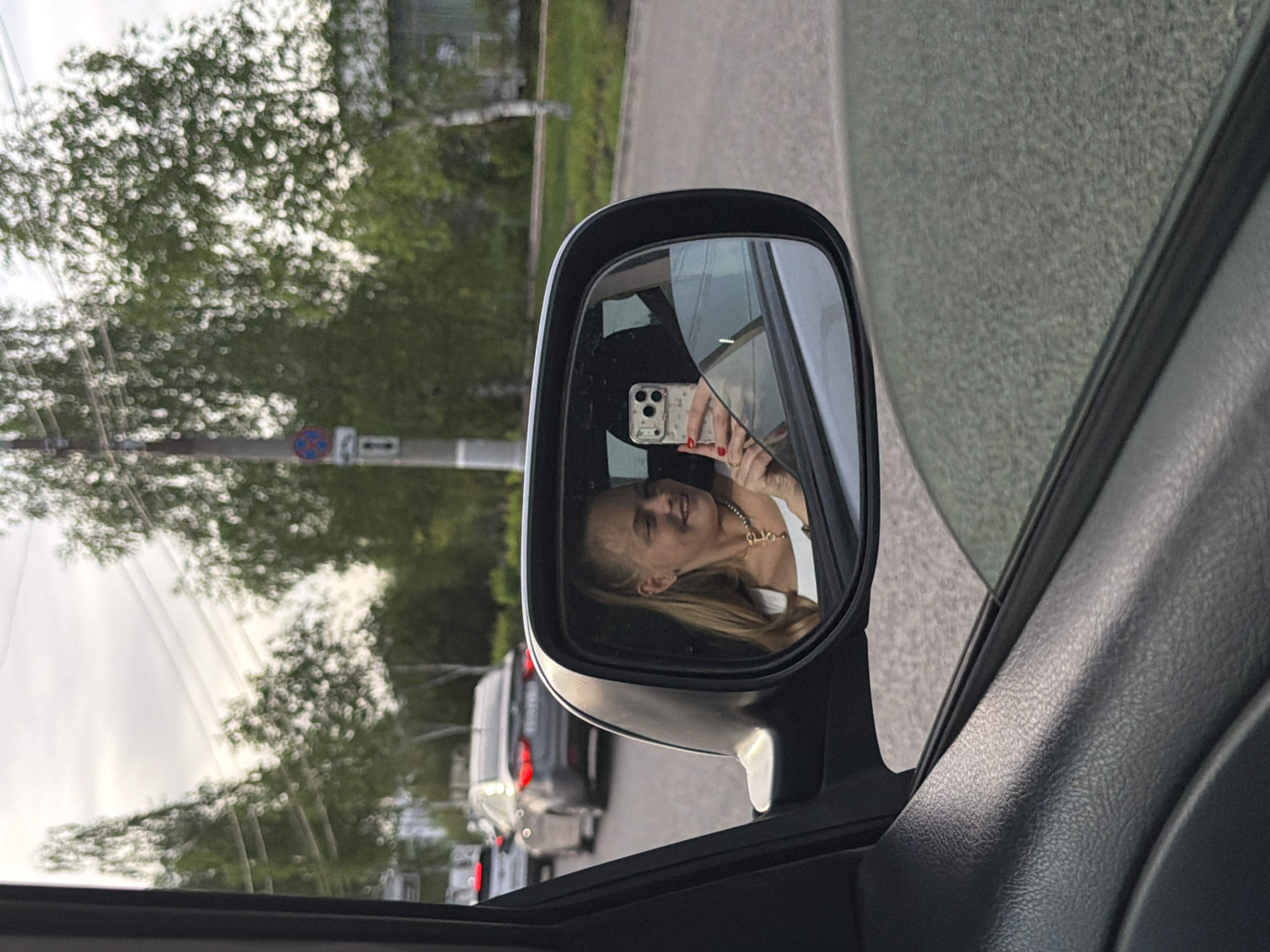 A view from inside a car shows a woman reflected in the car's side mirror, holding a smartphone in her right hand. She has long blonde hair and wears a white shirt and a gold chain. The side mirror shows the road behind the car with other vehicles. Outside the mirror, trees and a street signpost are visible, indicating a location within Izhevsk, Russia.