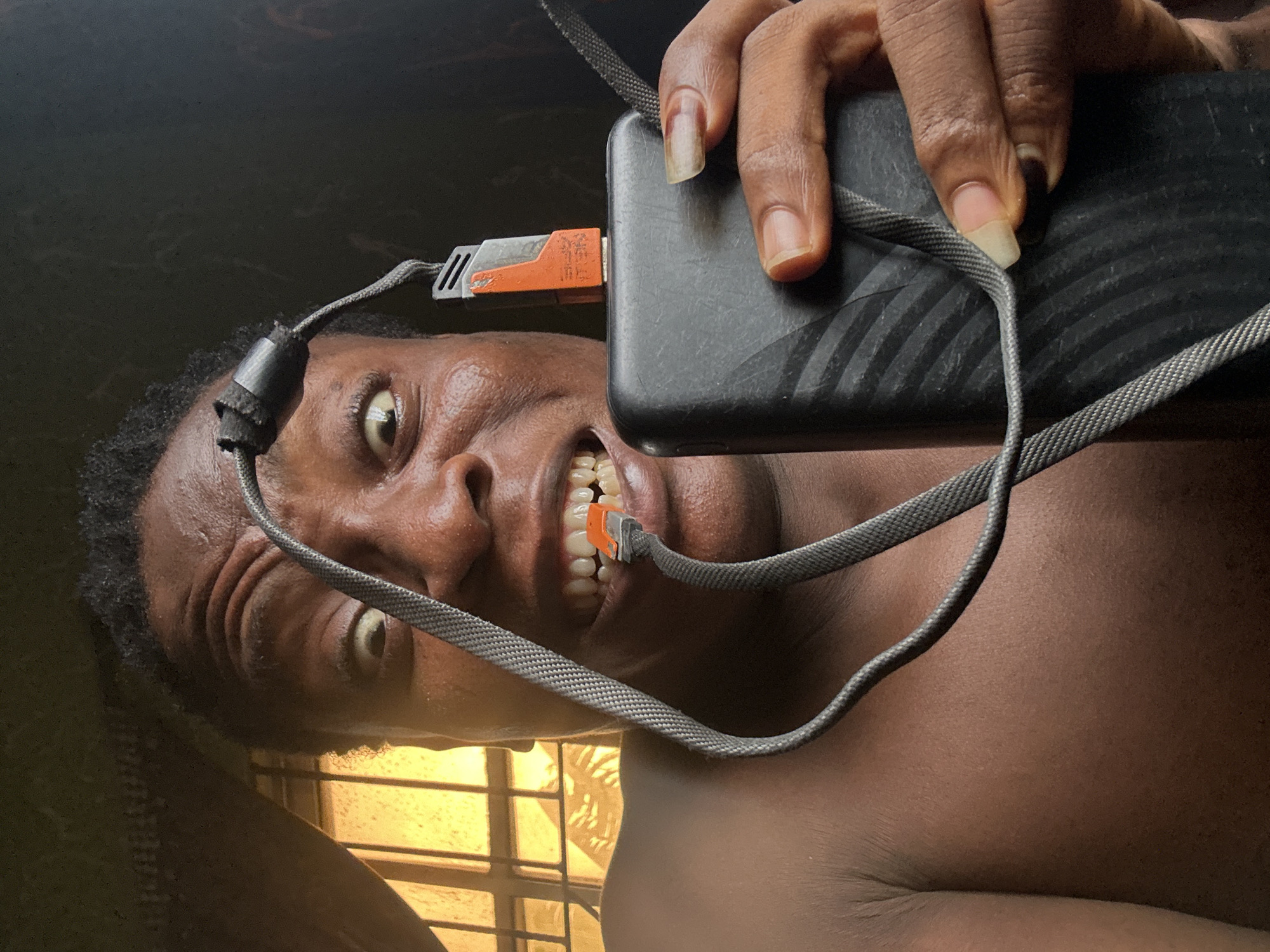A medium shot depicts an individual holding a black smartphone. The phone is connected to a charging cable, with one end of the cable in the person's mouth. The individual has dark hair and skin, with visible teeth. The background suggests a room with a window. The city and specific location are unknown.