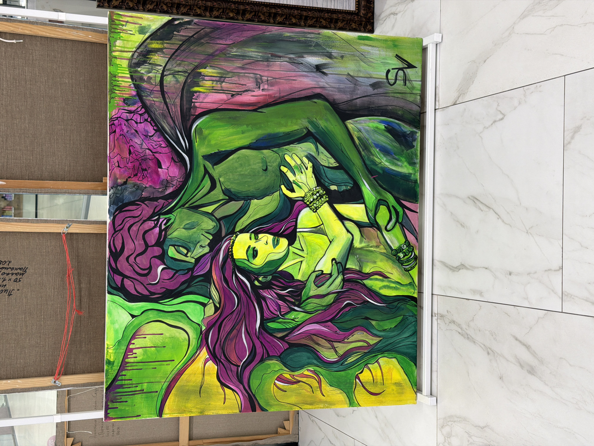 A large, vibrantly colored painting is displayed on a white easel. The painting, rendered in an expressive, abstract style, depicts two figures embracing. The dominant colors are shades of green, yellow, and purple, with black and pink accents. The figures have flowing hair and are depicted in a stylized manner. One figure appears to be larger and more muscular, embracing a smaller, more delicate figure. The background is a wash of green and yellow, with dripped paint effects in pink and purple. The painting is signed in the bottom right corner with the initials "SV." The easel is positioned on a light-colored tiled floor with a marble pattern. In the background, other artworks are visible on easels, and some of the canvases have labels and writing on them. The overall setting suggests an art exhibition or gallery space. There is no specific information in the image to indicate the location as Minsk, Belarus.