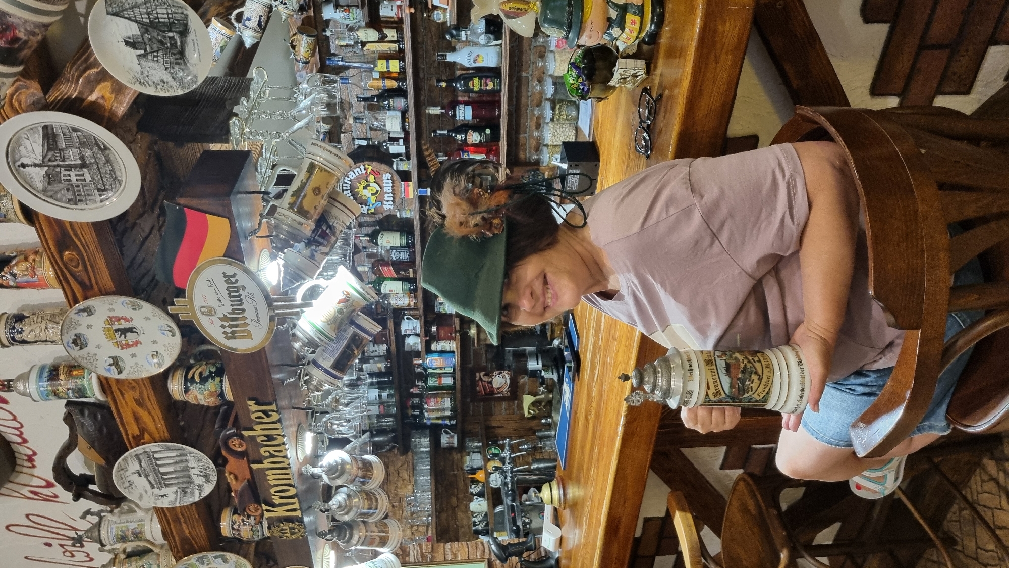 This is a medium shot, eye-level, interior photograph of a smiling woman sitting at a wooden bar, holding a large decorative beer stein. She is wearing a green hat with feathers and a light purple t-shirt, and denim shorts. The bar is lined with shelves displaying various beer steins, bottles of alcohol, and other German-themed decorations. The background shows a brick wall and more shelves filled with bottles. The scene appears to be in a German-themed bar or restaurant. The location of Chernivtsi, Ukraine, is not visible in the image.