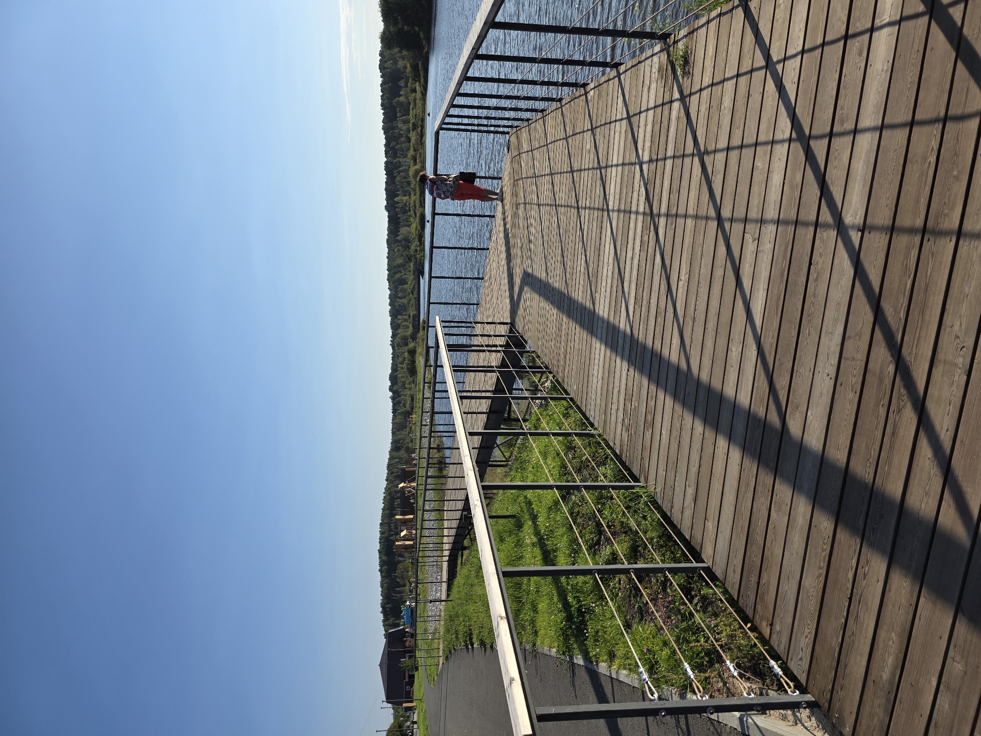 The image depicts a wooden boardwalk extending over a body of water, likely a lake or river, in the city of Miass, Russia. The sky is a clear, bright blue, indicating a sunny day. On the boardwalk, a single individual, a person wearing orange pants and a patterned shirt, stands near the edge, facing the water. The boardwalk is constructed of wooden planks and has a metal railing with rope infills. To the left of the boardwalk is a paved pathway, bordered by green grass and some vegetation. In the background, a tree line borders the water, and further in the distance, there are some structures, possibly part of a park or recreational area, including wooden playground equipment. The sunlight casts distinct shadows of the railing onto the boardwalk.