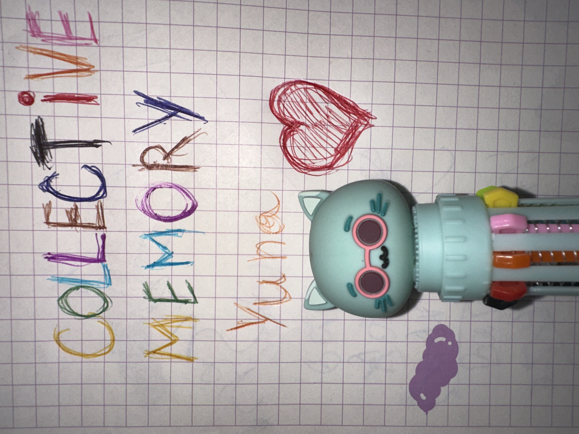 A multi-colored pen with a whimsical cat-like design rests on a piece of white graph paper, covered in purple grid lines. The image is a close-up, taken from a top-down perspective, showcasing a colorful and creative activity.
The pen, which has a light blue, cartoonish head resembling a cat with pink, circular glasses, is positioned horizontally across the paper. Its multi-color ink cartridges are visible at the end, displaying an array of vibrant hues, including pink, orange, red, black, purple, brown, blue, light blue, green, and yellow, indicating a diverse selection for writing and drawing.
On the graph paper, two words are written vertically in large, block letters, each letter in a different color. The word "COLLECTIVE" is spelled out with letters in yellow, green, light blue, purple, black, red, orange, pink, orange, and pink. Adjacent to it, the word "MEMORY" is written using yellow, green, light blue, purple, brown, and dark blue inks. Horizontally, to the right of these words, the name "Yume" is written in an orange or light brown script.
A red heart, filled with scribbled lines, is drawn near the top right of the paper. Towards the bottom right, a small, irregular purple blob or scribble is also visible, possibly a test mark from the pen. The overall scene suggests a moment of creative expression or note-taking. The setting is indoors, with the location context provided as Esenyurt, Turkey.