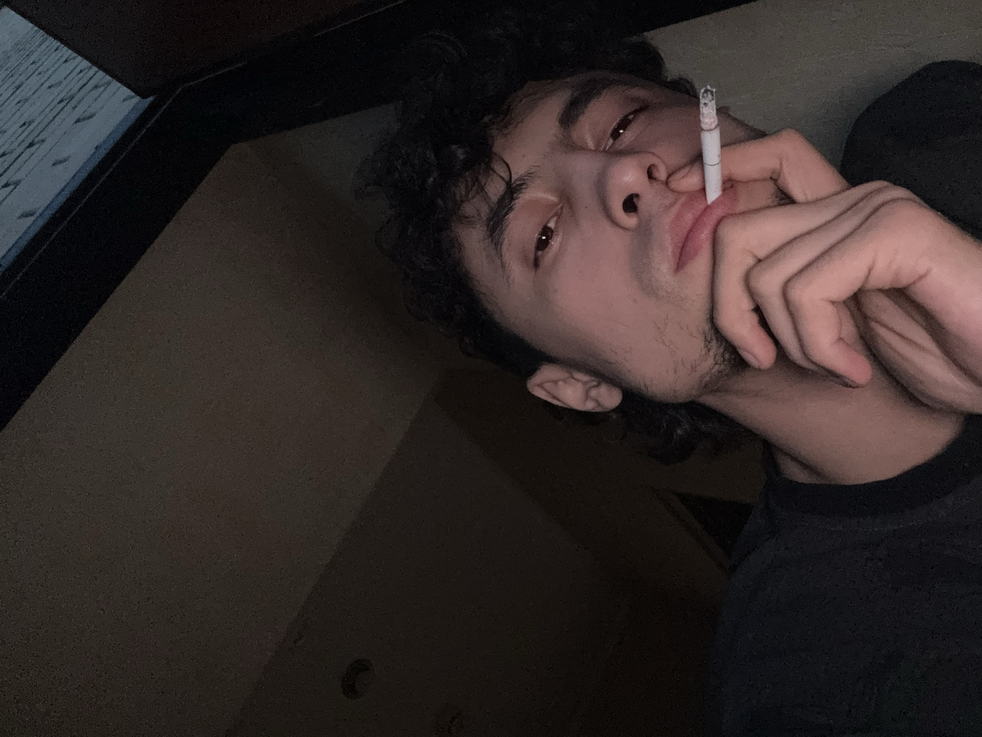 A close-up, low-angle shot shows a young man holding a lit cigarette to his lips, looking towards the camera. He has dark, curly hair and is wearing a dark t-shirt. His right hand is raised to his face, with his fingers supporting the cigarette. The background is dark and out of focus, with hints of what appears to be an interior space and possibly a window or doorway with a textured surface visible. The lighting is dim, with a warm cast.