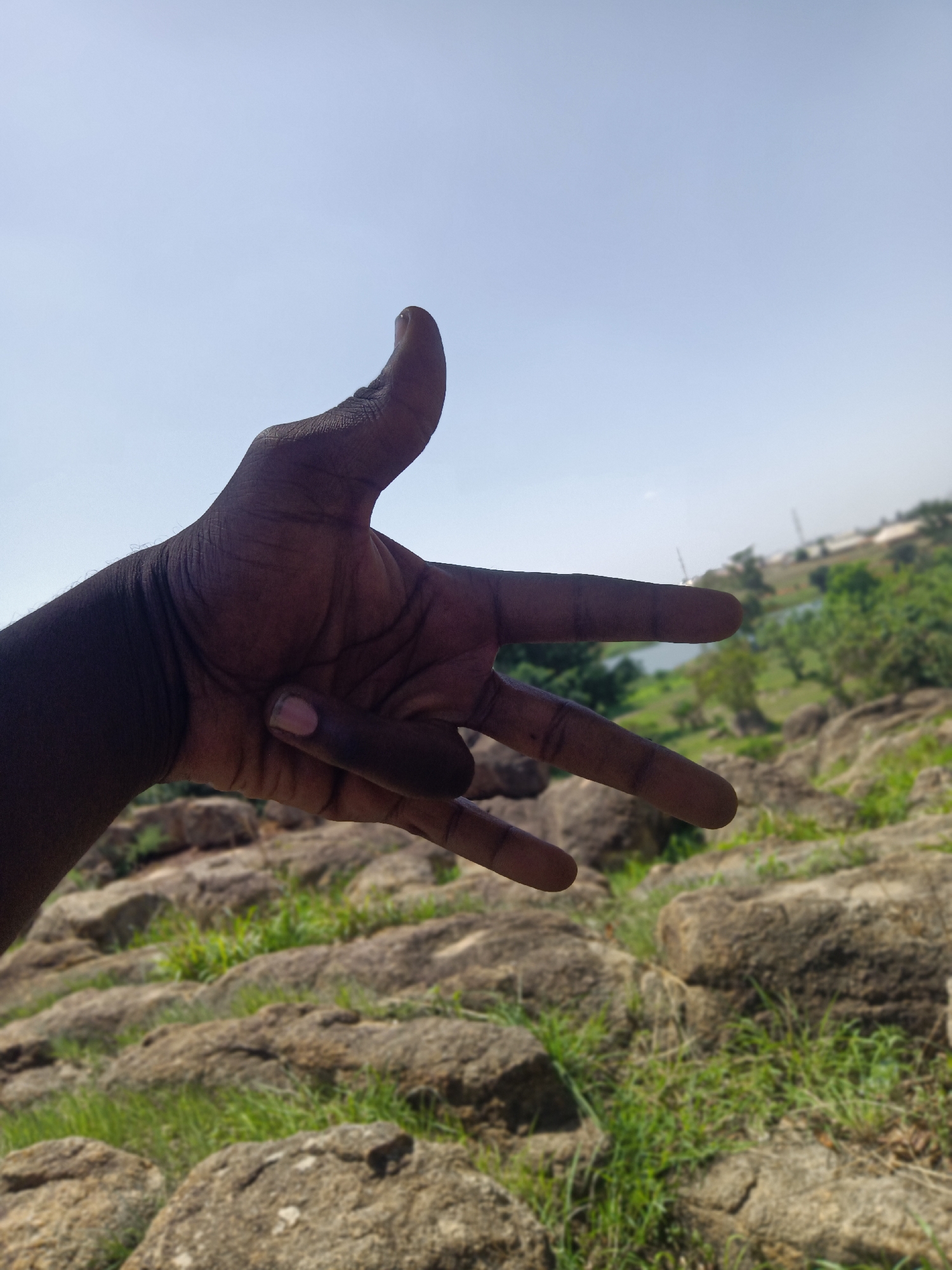 A dark-skinned person's hand is outstretched in front of a rocky, grassy terrain under a clear blue sky. The hand is positioned with the thumb extended upwards and the index, middle, and ring fingers extended outwards, while the pinky finger is tucked down. The background includes a body of water and distant structures, suggesting a location in Du, Nigeria, known for its natural rock formations and scenic landscapes. The environment appears natural and somewhat rural, with scattered vegetation among the rocks.