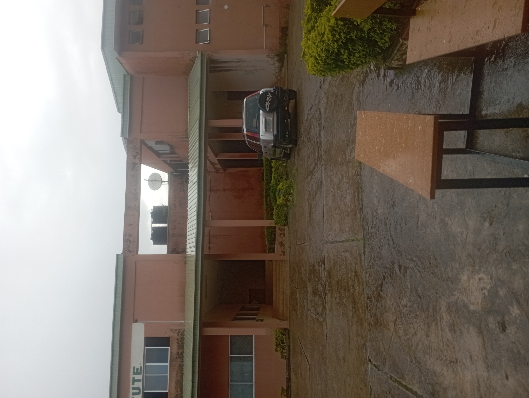 The image depicts the exterior of a light brown, two-story building in Jos, Nigeria, under an overcast sky. The building features a green metal roof and several windows, some of which are open. A covered walkway with pillars extends from the building to an entrance with steps. On the roof, two large black water tanks are visible, along with a satellite dish on a smaller structure.
In the foreground, a paved courtyard appears wet, suggesting recent rain. Two empty wooden benches with black metal legs are situated in the courtyard. A silver Honda CR-V SUV, with a spare tire mounted on the back, is parked to the right of the entrance. Dense green hedges line the sides of the building and the courtyard. The overall scene suggests a quiet, possibly institutional setting, likely a school or office building, after a rainfall in Jos.