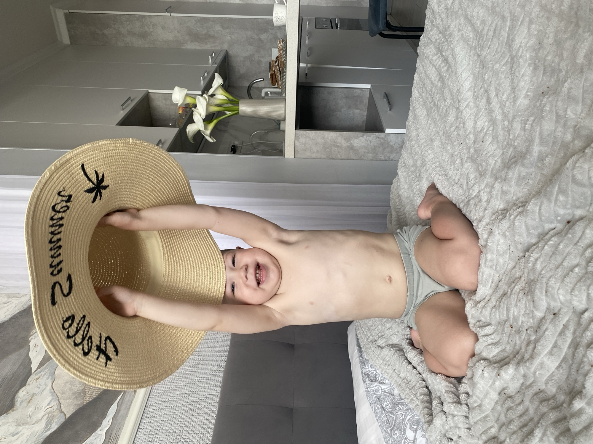A young boy is lying on a bed, holding up a straw hat. The hat has black text and a palm tree symbol on it. The boy is smiling and appears to be happy and playful. He is wearing light gray underwear. The bed is covered with a soft, textured white blanket. In the background, there are kitchen cabinets and a vase with white calla lilies. The overall setting suggests a home environment, possibly in Sochi, Russia, given the location context. The lighting suggests it is daytime, and there are no indications of weather conditions.