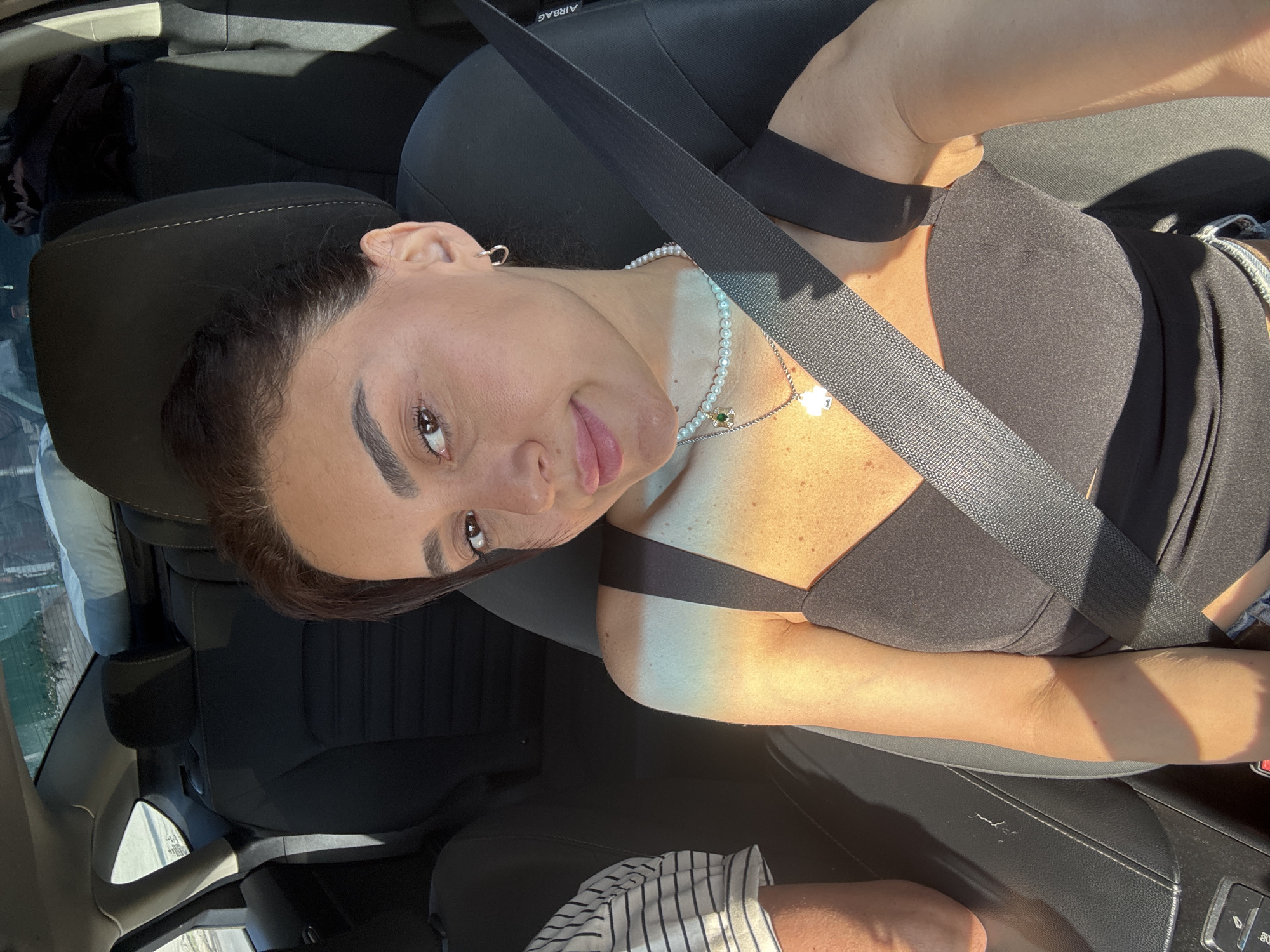 A woman with dark hair and brown eyes is sitting in the driver's seat of a car. She is wearing a black top and a pearl necklace. The car is parked, and sunlight is streaming in through the window, casting a warm glow on her skin. In the background, the seats of the car are visible, as well as a portion of the exterior landscape. The overall impression is one of a candid moment, possibly taken during a drive or a break on a sunny day in Tbilisi, Georgia.