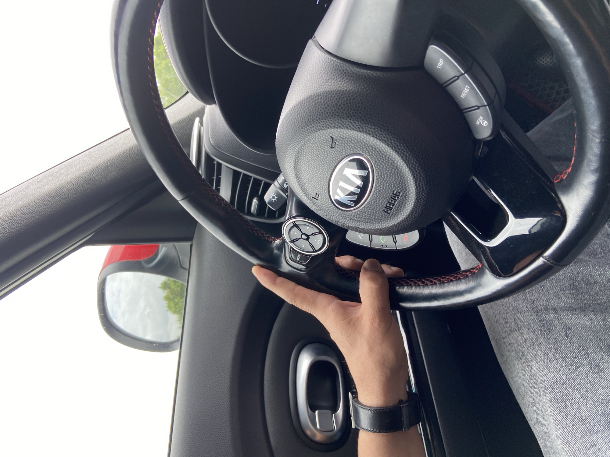 In Pervomaiskoe, Russia, a person's hand with a black wristwatch is seen resting on the steering wheel of a car. The steering wheel has red stitching and the Kia logo is visible in the center. A portion of the car's interior, including the door handle and dashboard, is also visible, along with a side mirror showing a cloudy sky and trees. The person appears to be driving, or about to drive, the vehicle.