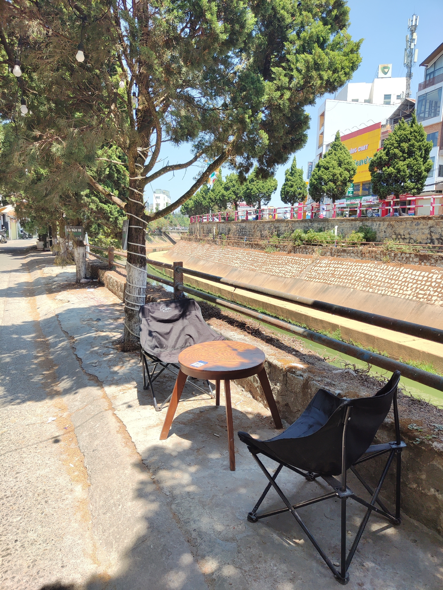 The image depicts an outdoor seating arrangement along a canal in what appears to be Da Lat, Vietnam, based on the architectural style of the buildings visible in the background. Two black, folding chairs are positioned on a concrete sidewalk beside a small, round, wooden table with three legs. The table sits between the chairs. A tree stands beside one of the chairs. The tree trunk is wrapped with a white material. A railing separates the sidewalk from the canal, which is lined with stone. The canal's water appears murky and greenish. In the background, buildings are visible, some with colorful signage. The sky is clear and blue. The scene is bathed in sunlight. A tree provides shade over the seating arrangement.