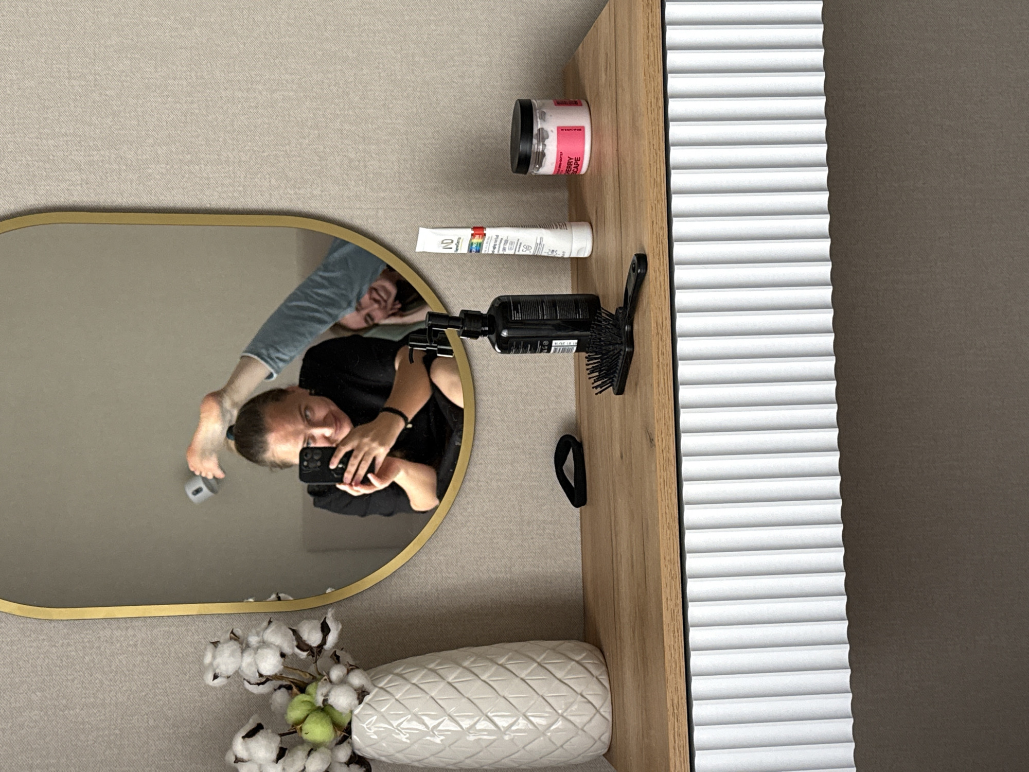A selfie of a person reflected in an oval mirror is captured indoors. The person is holding a smartphone, with their reflection showing their face and upper body, as well as their feet and legs positioned above their head. A second person is also partially visible in the mirror's reflection, appearing upside down relative to the primary subject.
Adjacent to the mirror on a wooden surface are an array of personal care items. These include a jar of a berry-themed product, a tube of ND brand cream, and a black hair dryer with a brush attachment. A black hair tie is also present.
A white vase filled with cotton flower stems sits beneath the mirror. The background consists of a white ridged surface and a neutral colored wall. There is no explicit evidence indicating that the location is in Izhevsk, Russia.
