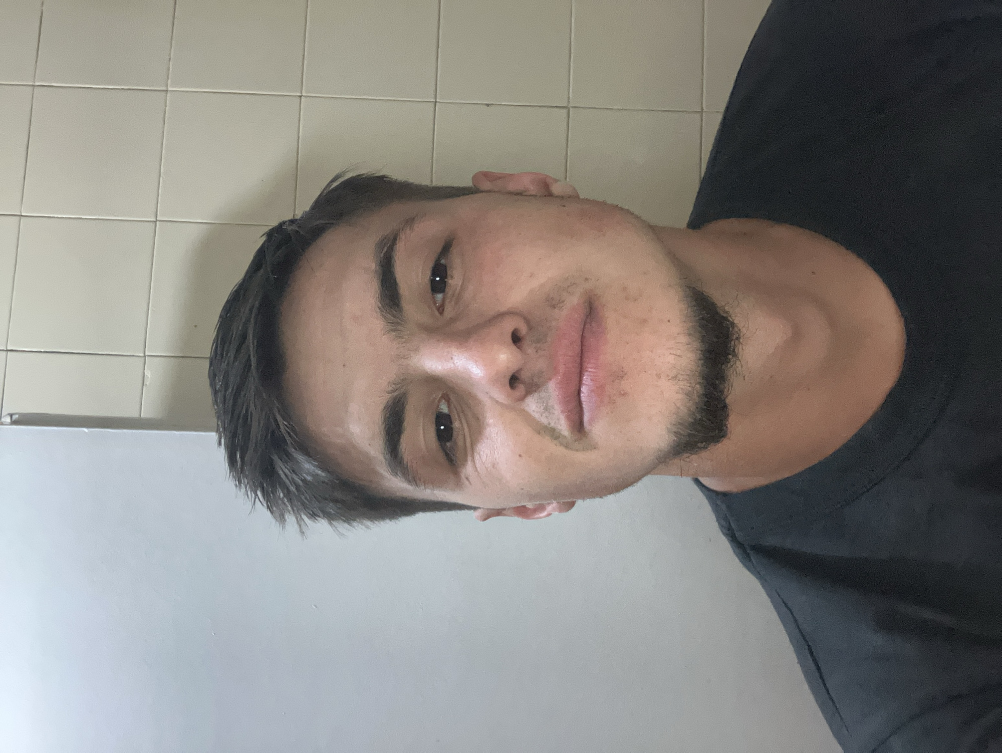 A close-up headshot of a man with short, dark hair and a trimmed beard. He is wearing a dark t-shirt. The background is a tiled wall, possibly in a bathroom, and a plain, light-colored wall. The location in Plovdiv, Bulgaria is not discernible from the image.