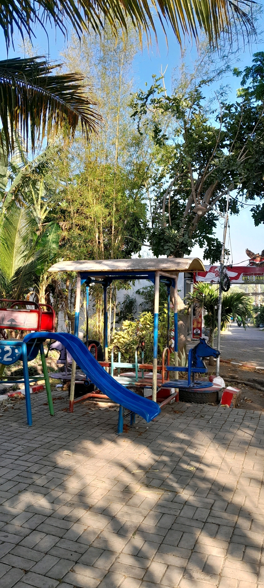 The image depicts an outdoor playground scene with a blue slide as the central focus. The playground structure is made of metal with blue and red accents. A blue horse-shaped rocker is attached to one side of the structure. The ground is paved with interlocking gray bricks, and shadows from surrounding trees are cast across the paving. In the background, there are various types of vegetation including tall bamboo-like plants and leafy trees. A red and white banner is visible in the far background, and a street or pathway leads away from the playground. The sky is clear and blue. The location is identified as Bendungan, Indonesia. There are no people visible in the image.
