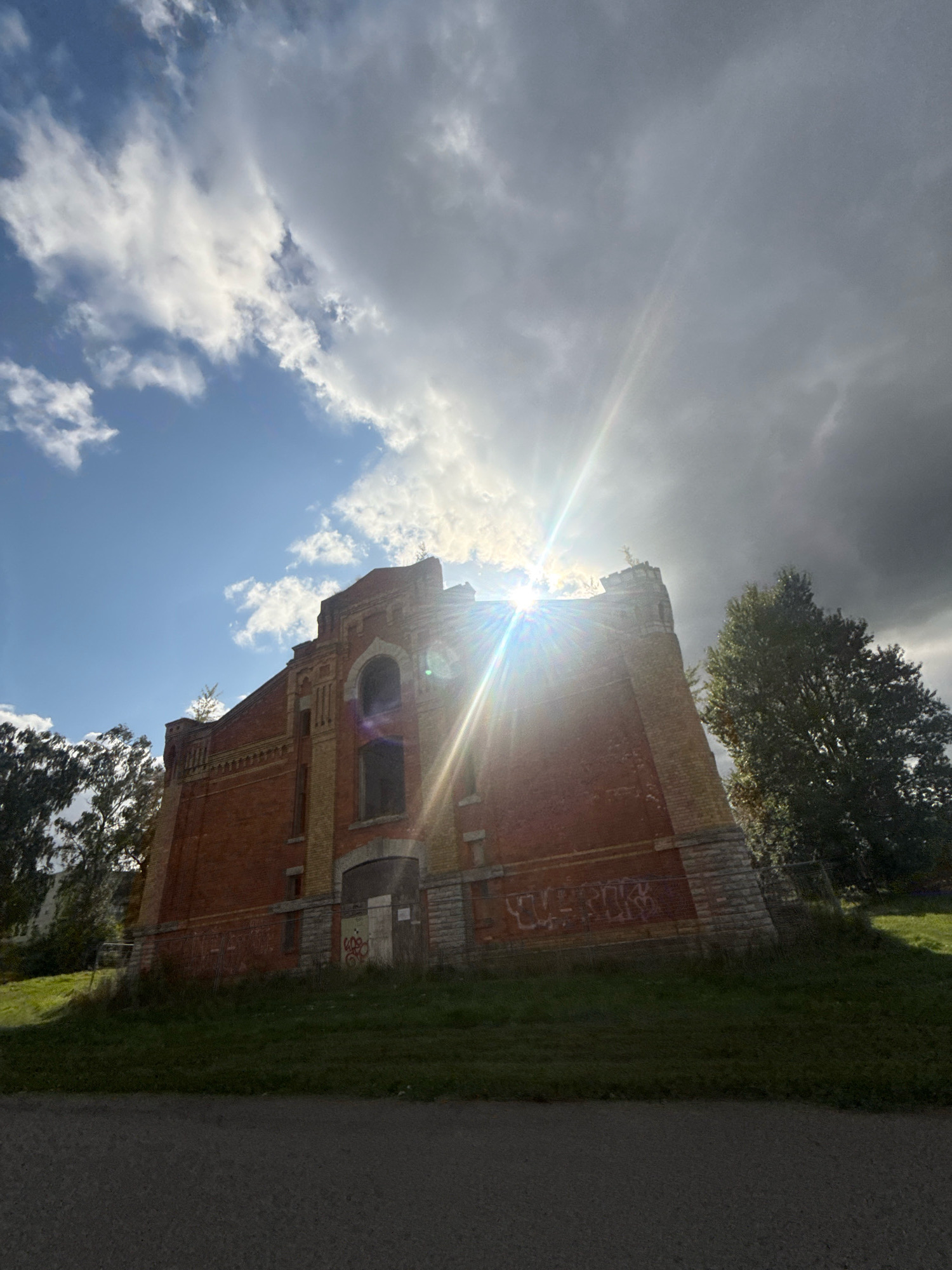 The image captures a brick building with yellow accents, likely an abandoned or historical structure, situated in Tallinn, Estonia. The building has arched windows, some boarded up, and graffiti on its lower sections. It stands on a grassy incline, with trees to its left and right. The sky is a mix of bright blue with scattered white clouds and darker, more ominous clouds. Sunlight streams through the clouds, creating lens flare and illuminating parts of the building and the sky. A paved path is visible in the foreground.