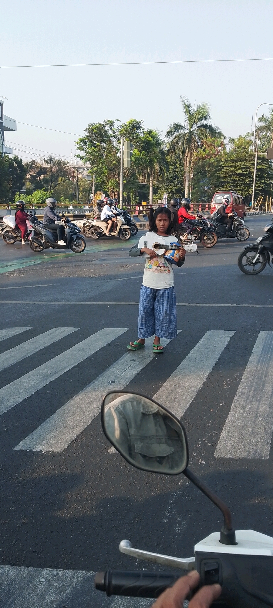 A young Indonesian girl with pigtails is standing on a crosswalk in Bekasi, Indonesia, holding a ukulele. She is wearing a white t-shirt with a colorful cartoon image and loose blue plaid pants. Green flip-flops are on her feet. She is looking directly at the camera. Behind her, traffic flows, with several motorcycles and a red van visible. The scene takes place on a street with a crosswalk marked by white stripes. The image is taken from the perspective of someone on a motorcycle, with the handlebars and a rearview mirror in the foreground. The sky is clear and bright.