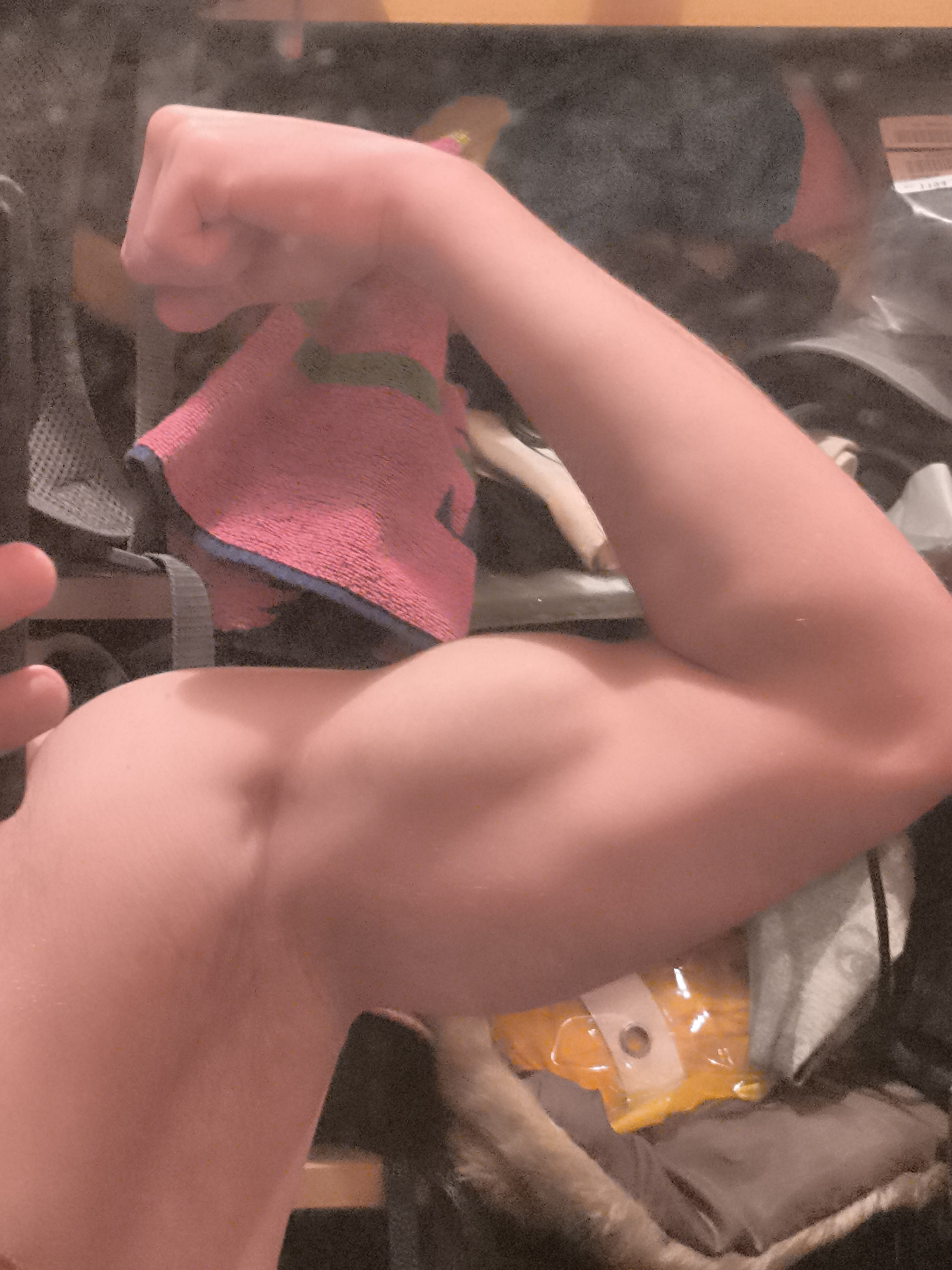The image displays a close-up shot of a person's light-skinned left arm, flexed to prominently show the bicep, with the hand clenched into a fist. A vibrant pink towel, featuring blue and green trim, is draped casually over the upper arm and shoulder region.
The scene is set indoors in Sofia, Bulgaria. The background is blurred and appears cluttered, suggesting an informal personal space such as a room, changing area, or perhaps a living space. Among the indistinct objects in the background, a grey mesh fabric, possibly part of a backpack or sports gear, can be discerned. In the lower right corner, a clear plastic bag containing orange or yellow contents, possibly food or a packaged item, is visible, secured with a white label or fastener.
The central activity is the individual flexing their arm, a common gesture to demonstrate physical strength or muscle tone. The lighting appears to be artificial and somewhat dim, contributing to a slightly grainy texture across the image. No specific time of day or weather conditions are discernible from this indoor setting, and no identifying location cues for Sofia are explicitly visible beyond the provided context. The person's face is not visible, so no specific emotions are conveyed, though the act of flexing a bicep often implies confidence or a playful display of strength. There is no visible text within the image.