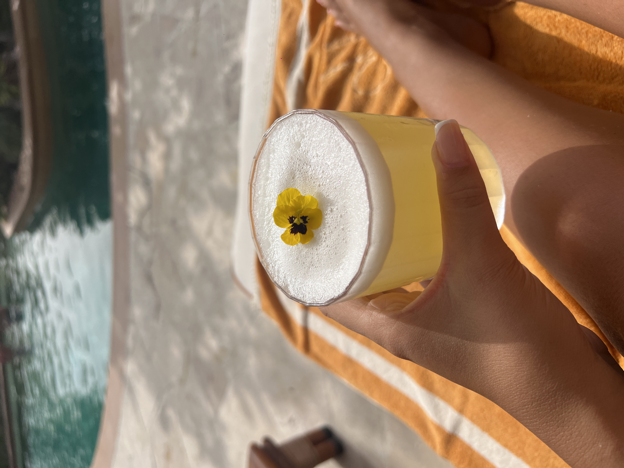 A high-angle shot shows a person holding a glass of yellow cocktail with a white foamy top garnished with a yellow flower. The person's hand and part of their leg are visible against a horizontally striped orange and white towel. To the left, out of focus, is a pool or body of water with a marble or tiled edge. The setting is outdoors, likely near a swimming pool. There is no specific information in the image that pinpoints the location to Dubai.