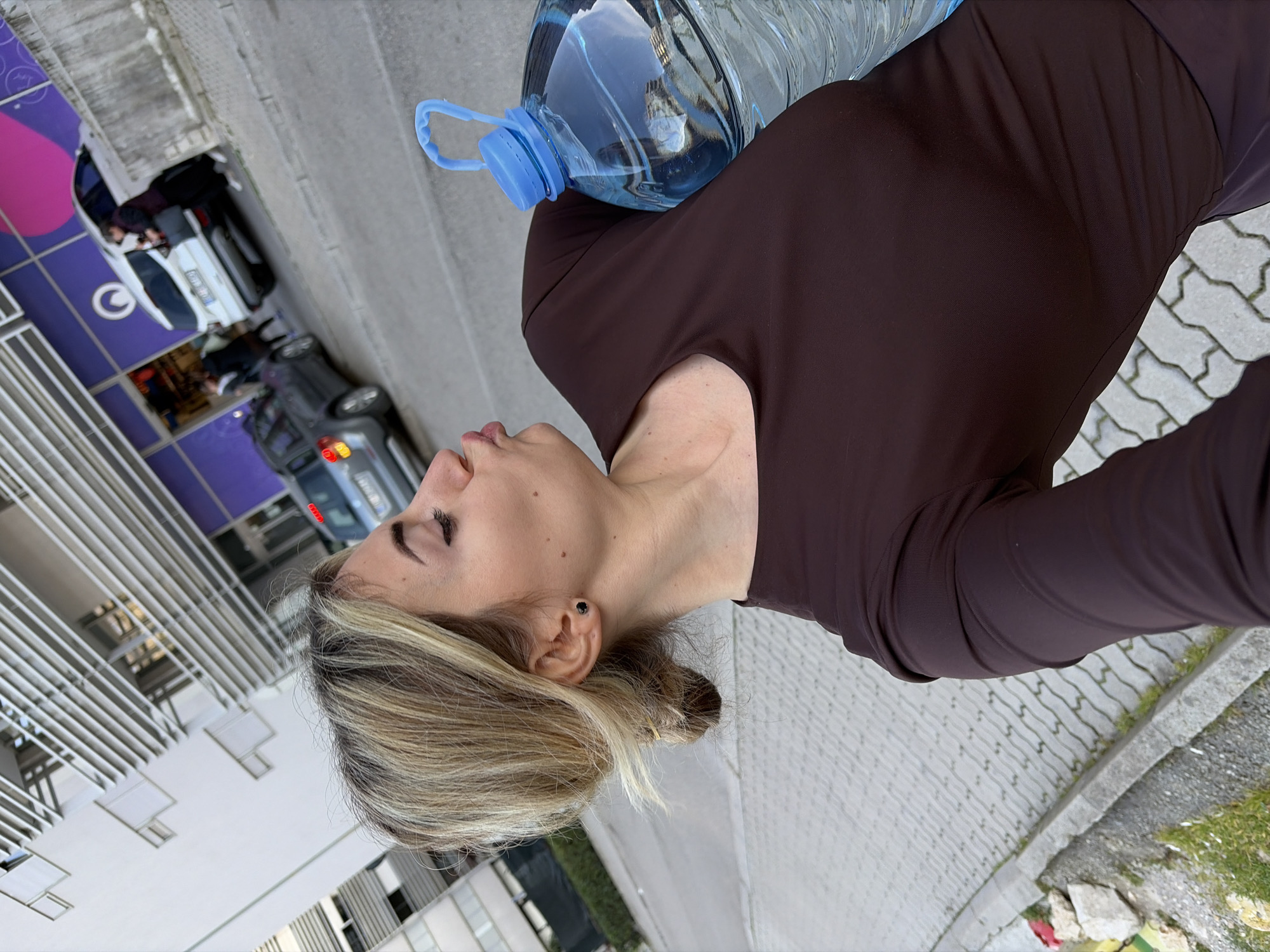 A woman with blonde hair, styled in a bun, is captured in a close-up, angled shot. She wears a dark brown long-sleeved top and a small black stud earring in her left ear. Her head is turned to her right, and her lips are pursed in a pouting expression. She holds a large, clear plastic water bottle with a blue handle and cap against her torso.
The scene is an urban outdoor environment, specifically identified as being near the Okova store at Cetinjski put bb, Budva, Montenegro. The foreground shows a sidewalk paved with grey interlocking pavers, bordered by a concrete curb with some scattered rocks and sparse grass.
Beyond the sidewalk, an asphalt street is visible. A grey hatchback car is parked on the street in the mid-ground. Further back, a white SUV is parked in front of a building with a distinctive purple facade, which features a large white "O" logo and abstract magenta designs. An open entrance to a commercial establishment is visible on this facade, with several blurred figures, appearing to be people, near the entrance and the white SUV. To the left of the purple-fronted building, a modern, multi-story white building with prominent horizontal architectural elements (louvers or balcony rails) is visible under a light sky.