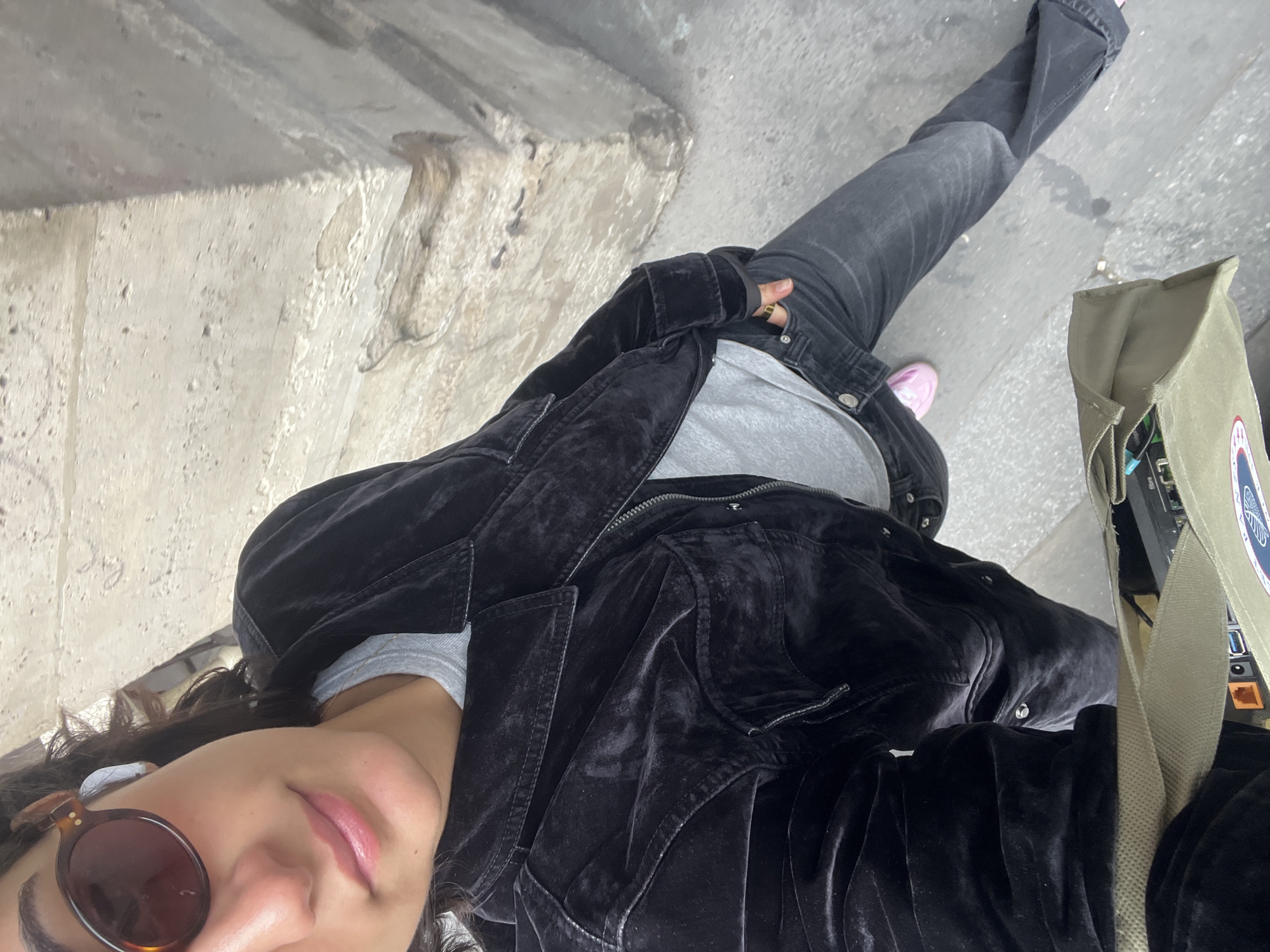 A high-angle, tilted selfie captures a person in an urban setting, likely Paris, France. The individual, seen from the chest upwards and including their lower body, has dark, curly hair and wears dark, round sunglasses with a tortoiseshell frame. Their expression is neutral. They are dressed in a grey t-shirt layered under an unzipped black velvet jacket and dark grey or black jeans. A gold-toned ring is visible on their right hand. One foot, clad in a bright pink shoe, is visible on the ground.
To the right of the person, a olive green canvas bag or tote bag is slung over their shoulder, revealing various electronic components, including cables, ports, and a circuit board or small device, protruding from its opening. An earbud is visible in the person's left ear. The background consists of a textured concrete or asphalt ground and a large, rough concrete wall or structure, possibly part of an underpass or bridge, suggesting an industrial or infrastructural environment. The overall lighting is soft, indicating daytime, possibly overcast. A circular emblem or patch with unreadable text is visible on the green bag.