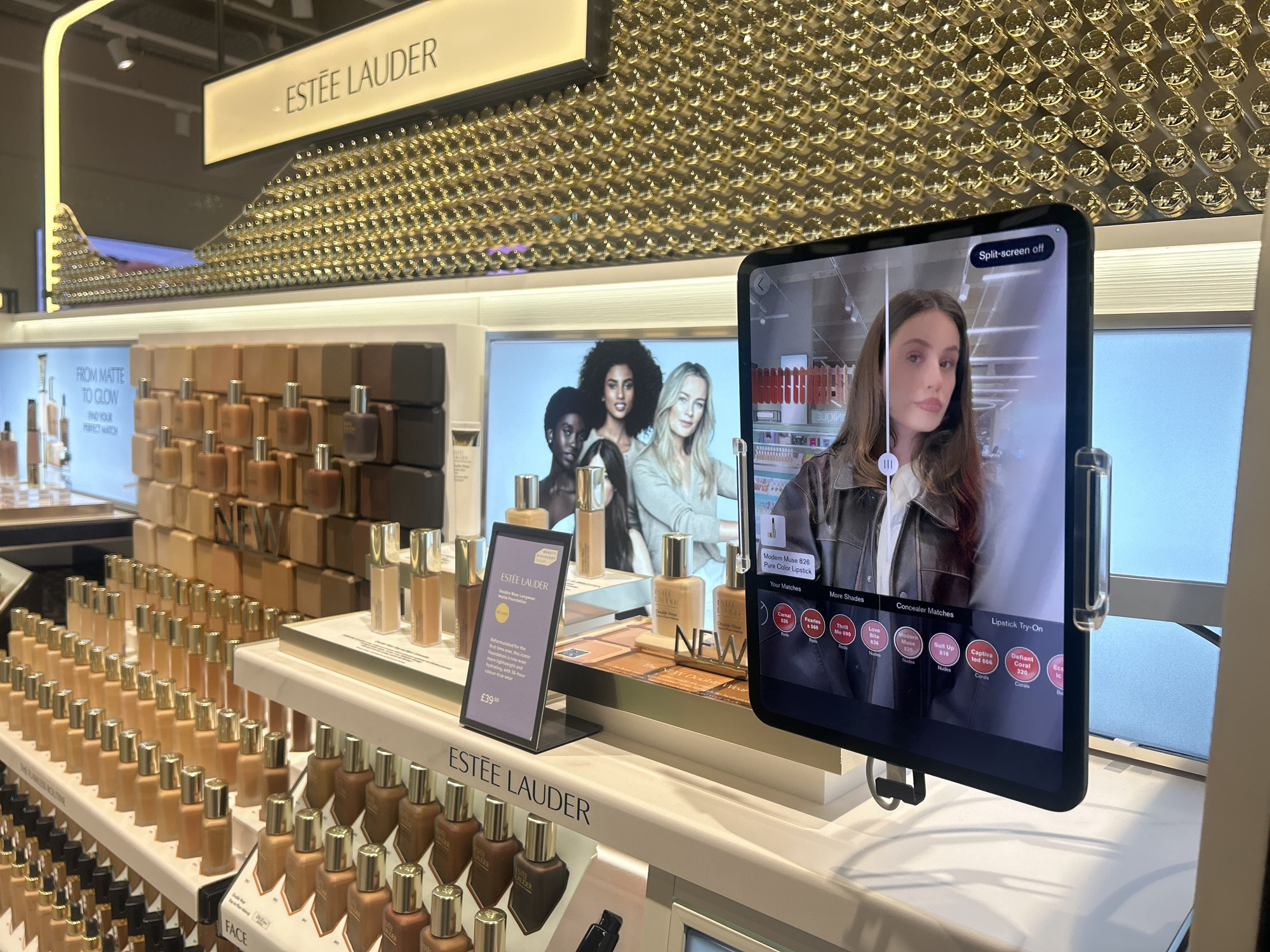 The image shows a makeup display, likely inside a retail store in the city of Bath, United Kingdom. The focus is on an Estée Lauder display featuring rows of foundation bottles of varying shades on tiered shelves. Behind the product display are advertisements featuring three diverse female models and an Estée Lauder product. In the foreground, there's a tablet-like device with a female model's face visible on screen. Buttons are visible on the sides of the screen. Also, on the table there is a price tag. The store environment is well-lit.