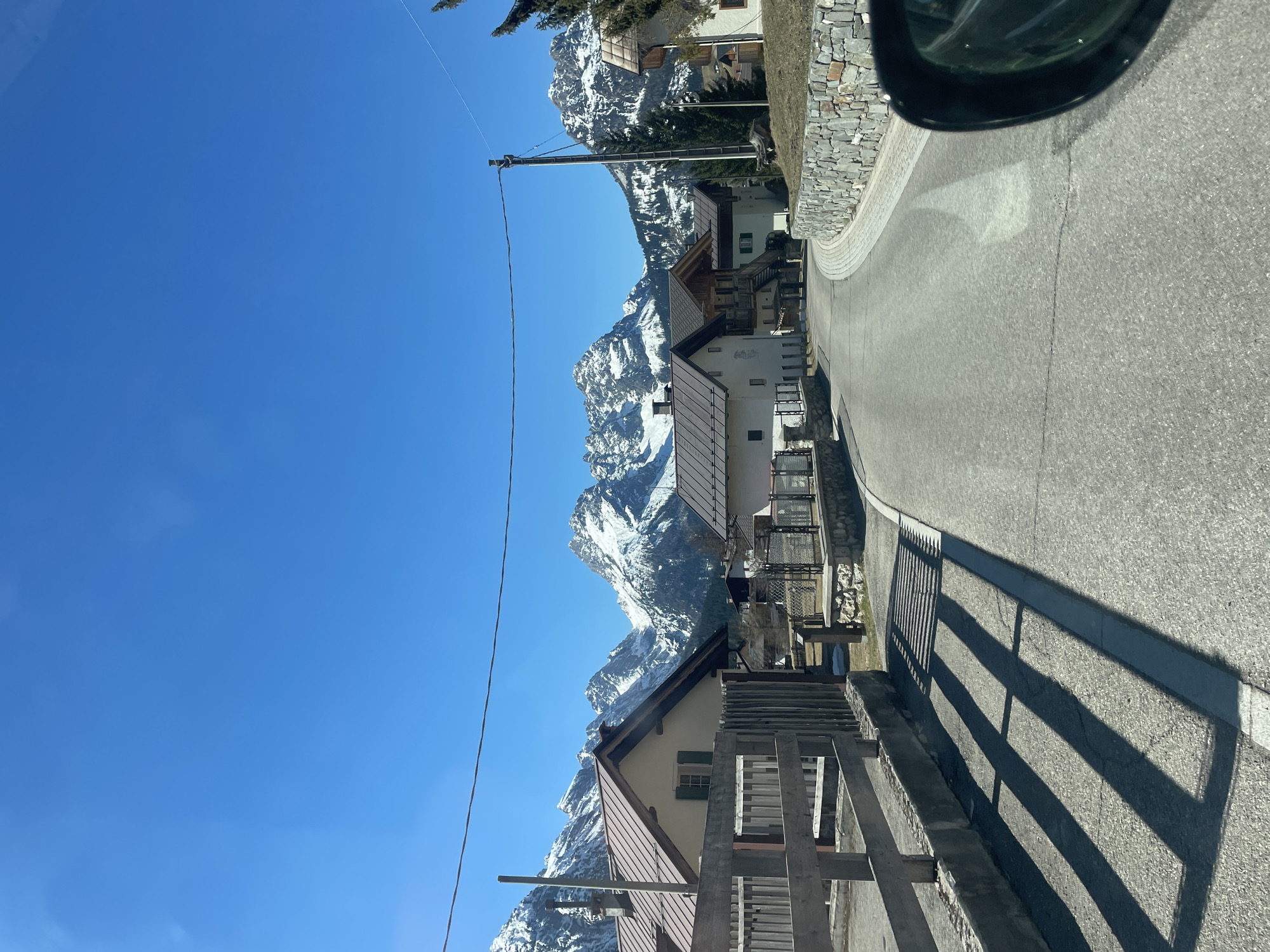 The image depicts a road winding through the town of Sappada, Italy, with snow-capped mountains in the background. The road is asphalt and appears well-maintained. Houses with traditional Alpine architecture are situated along the roadside. A utility pole with wires is visible against the clear blue sky. The sunlight casts shadows from a wooden fence onto the road surface. A portion of a car's side mirror is visible in the upper right corner of the image.