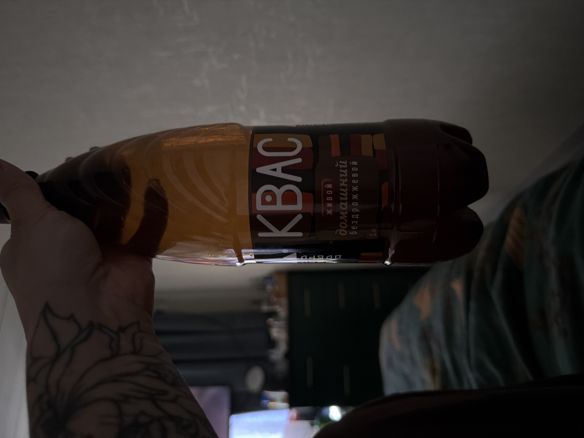 A person is holding a plastic bottle of "Kvas" drink. The label on the bottle reads "КВАС" in large white letters, with additional text in Russian indicating it is "live" and "homemade," and that it is yeast-free. The bottle is brown, and the liquid inside appears dark brown. The person holding the bottle has a tattoo on their arm, and they are in what seems to be a room with other objects and furniture visible in the background. Based on the label, the drink is likely from Russia. Given the Russian name on the bottle, the city of Sarapul, Russia could be a potential location.