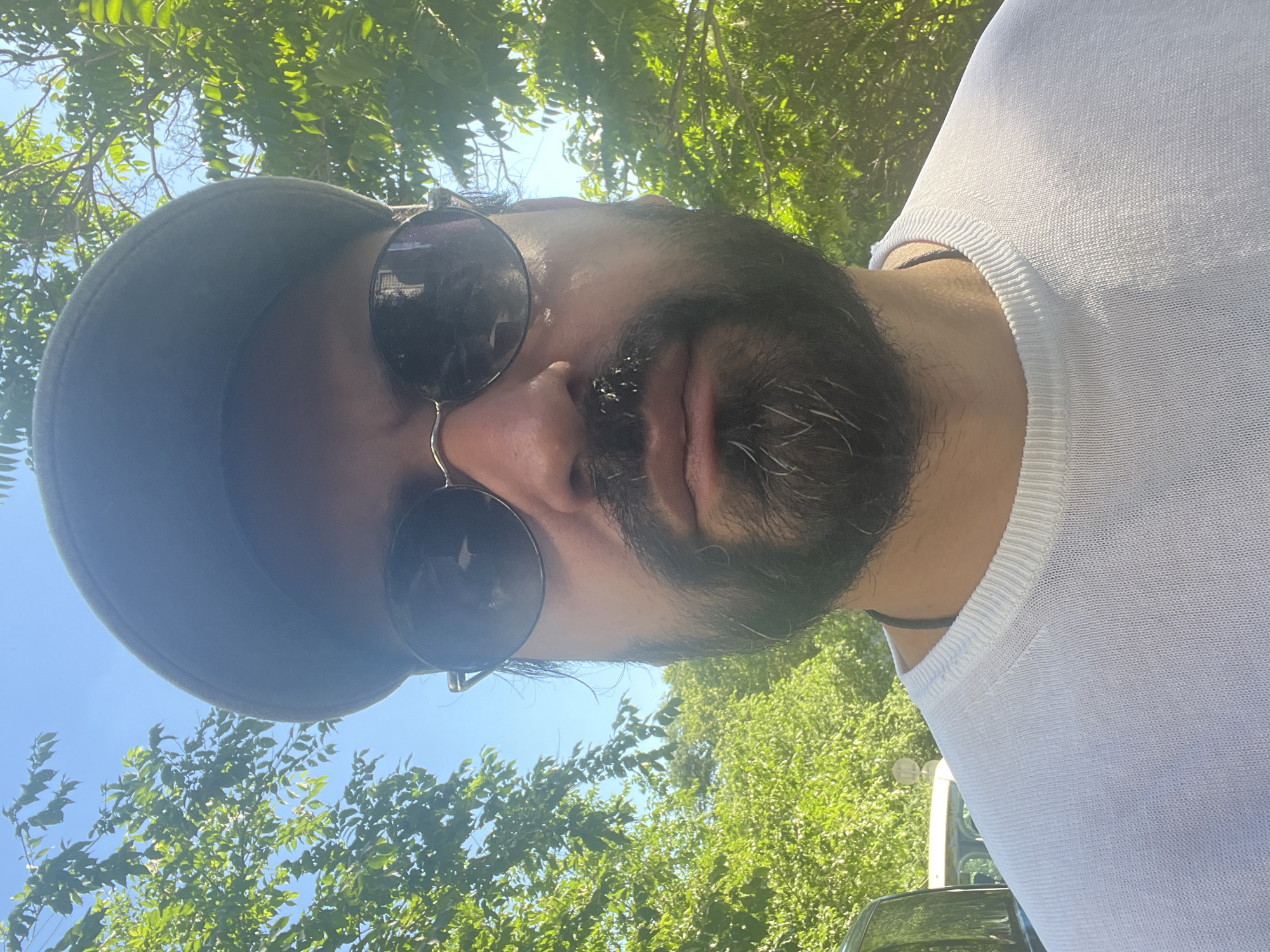 The image is a close-up, slightly tilted selfie of a person wearing a dark baseball cap and round sunglasses. The person has a beard and is looking towards the camera. They are wearing a white, knit shirt with a textured neckline. The background is filled with lush green foliage, suggesting an outdoor setting with trees. The bright sunlight and clear blue sky visible through the leaves indicate it is daytime and the weather is fair. There are no visible text elements in the image. The overall impression is casual and relaxed, possibly taken on a sunny day outdoors.