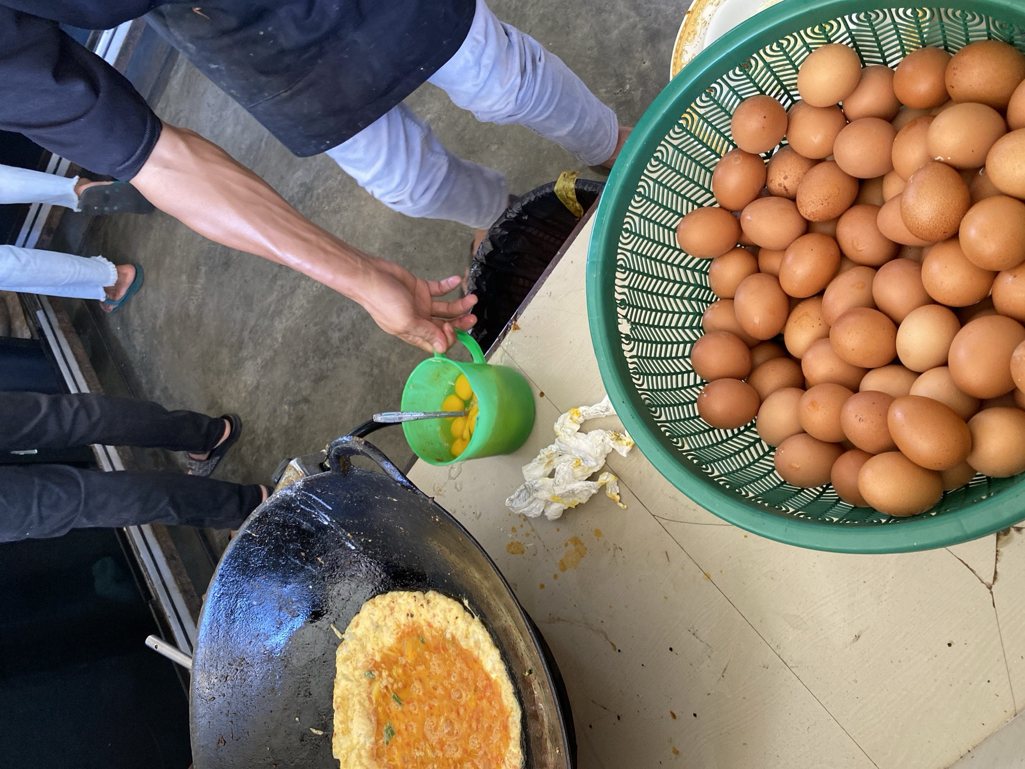 In Banda Aceh, Indonesia, a person's arm, glistening with sweat, is visible as they reach towards a green plastic container holding several cracked eggs. Adjacent to this is a large black wok containing a cooked omelet with a bright orange filling. On the table, a crumpled white paper towel stained with yellow is present, along with a green plastic basket filled with numerous brown eggs. In the background, the legs of two individuals are visible; one is wearing black pants and sandals, while the other is in light-colored pants and flip-flops. The setting appears to be an outdoor food stall or preparation area, with a concrete floor and a dark background structure.