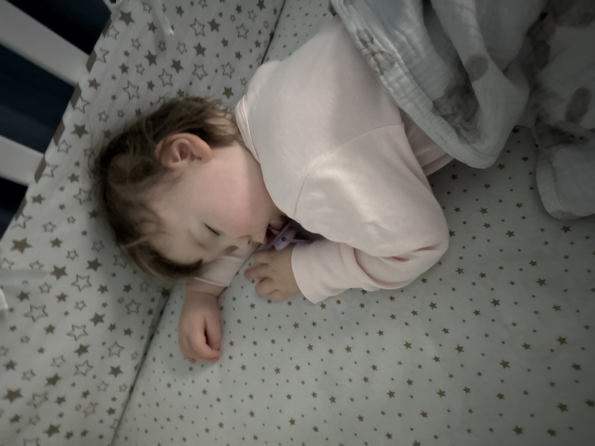 A young child sleeps on a star-patterned crib sheet, covered partially with a blanket. The child is lying on their side with a pacifier in their mouth. The crib has white railings. The location is not discernible as Moscow, Russia.