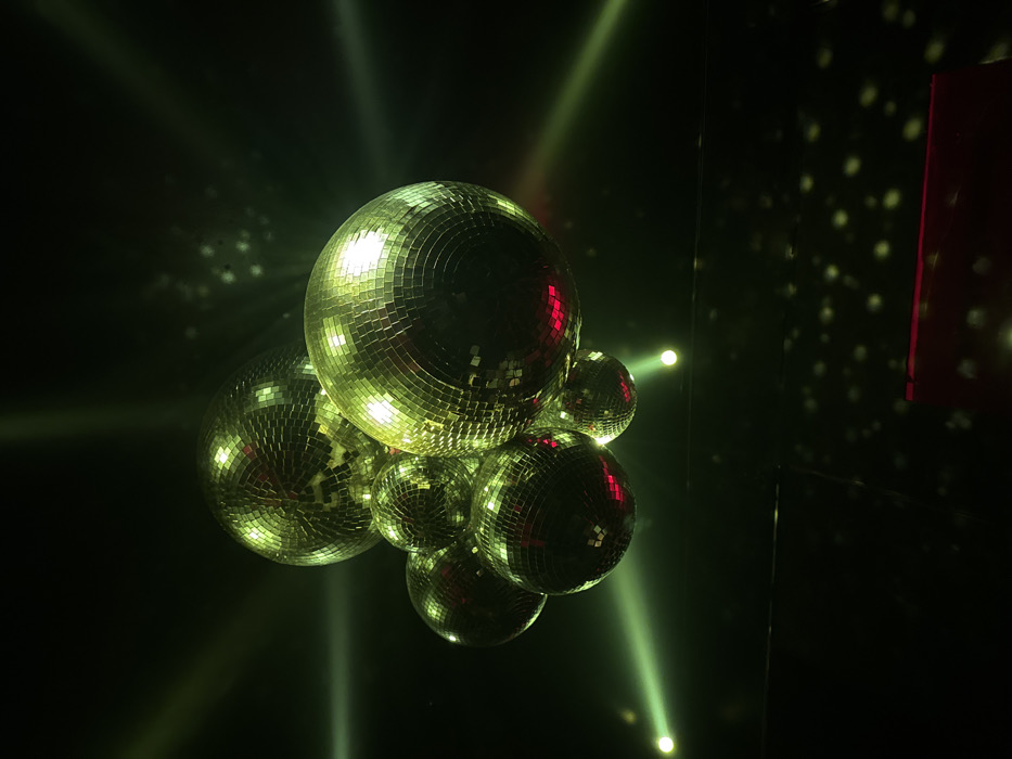 A cluster of approximately seven mirrored disco balls of various sizes hangs suspended in a dimly lit interior space, characteristic of a nightclub or event venue, located in Paris, France. The disco balls are composed of numerous small, square reflective facets. Strong beams of green-yellow light project from off-camera sources, illuminating the spheres and creating visible light rays through the dark environment. The facets of the disco balls reflect these lights, with distinct red reflections visible on the underside of several spheres. Scattered points of light, originating from the disco balls' reflections, are cast upon a dark surface in the lower right background. The overall scene is dark, ambient, and devoid of any visible individuals or specific human actions.