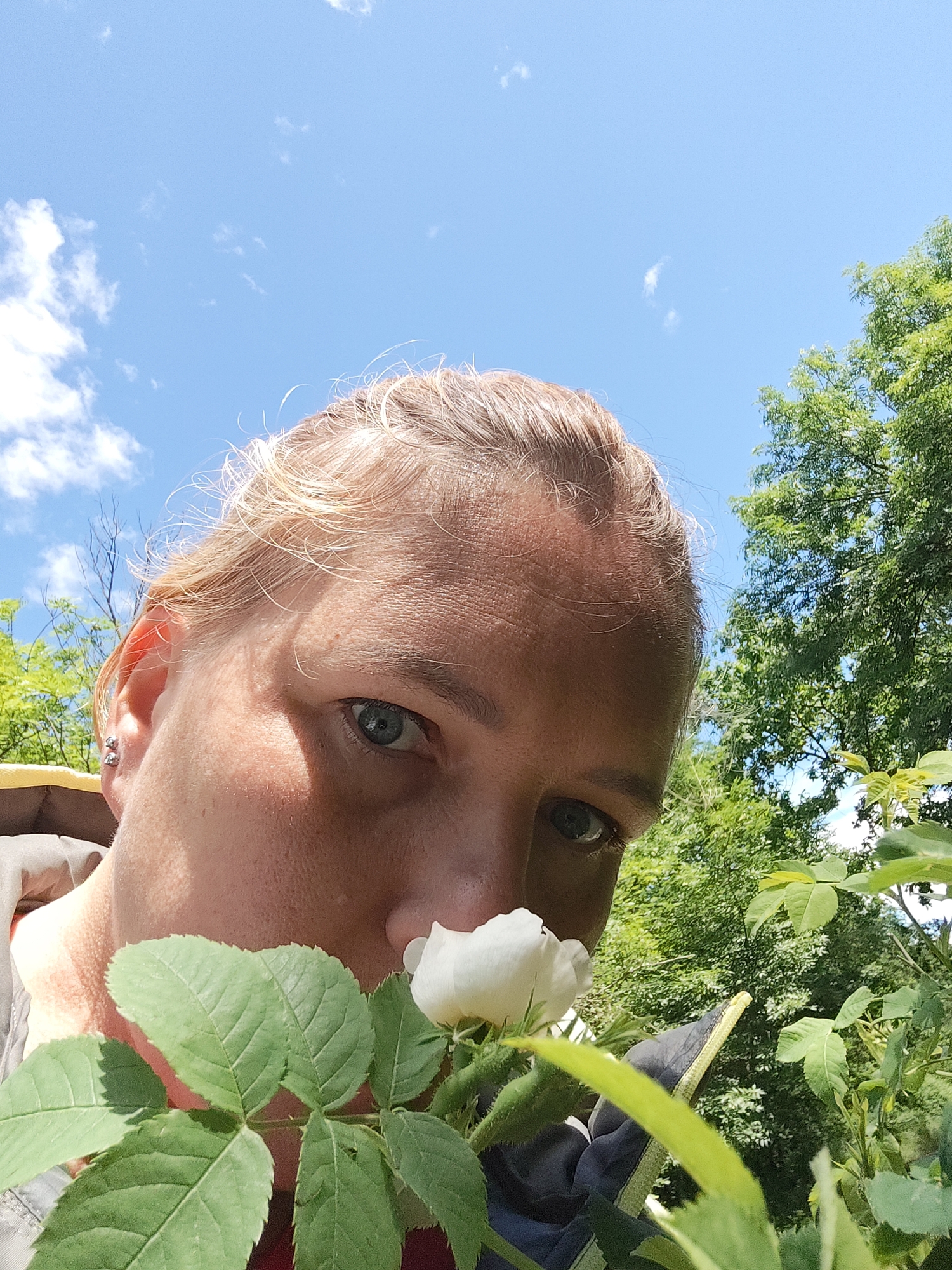 A close-up, low-angle shot features a fair-skinned person with light blonde hair and blue eyes, gazing directly at the camera. A white rose blossom is positioned near their nose. The background includes green foliage and a blue sky with scattered clouds. A portion of a yellow jacket is visible on the left. The scene appears to be set outdoors in a garden or natural area. While the setting is outdoors, the specific location within the city of Svobody, Russia cannot be determined from the visual data.