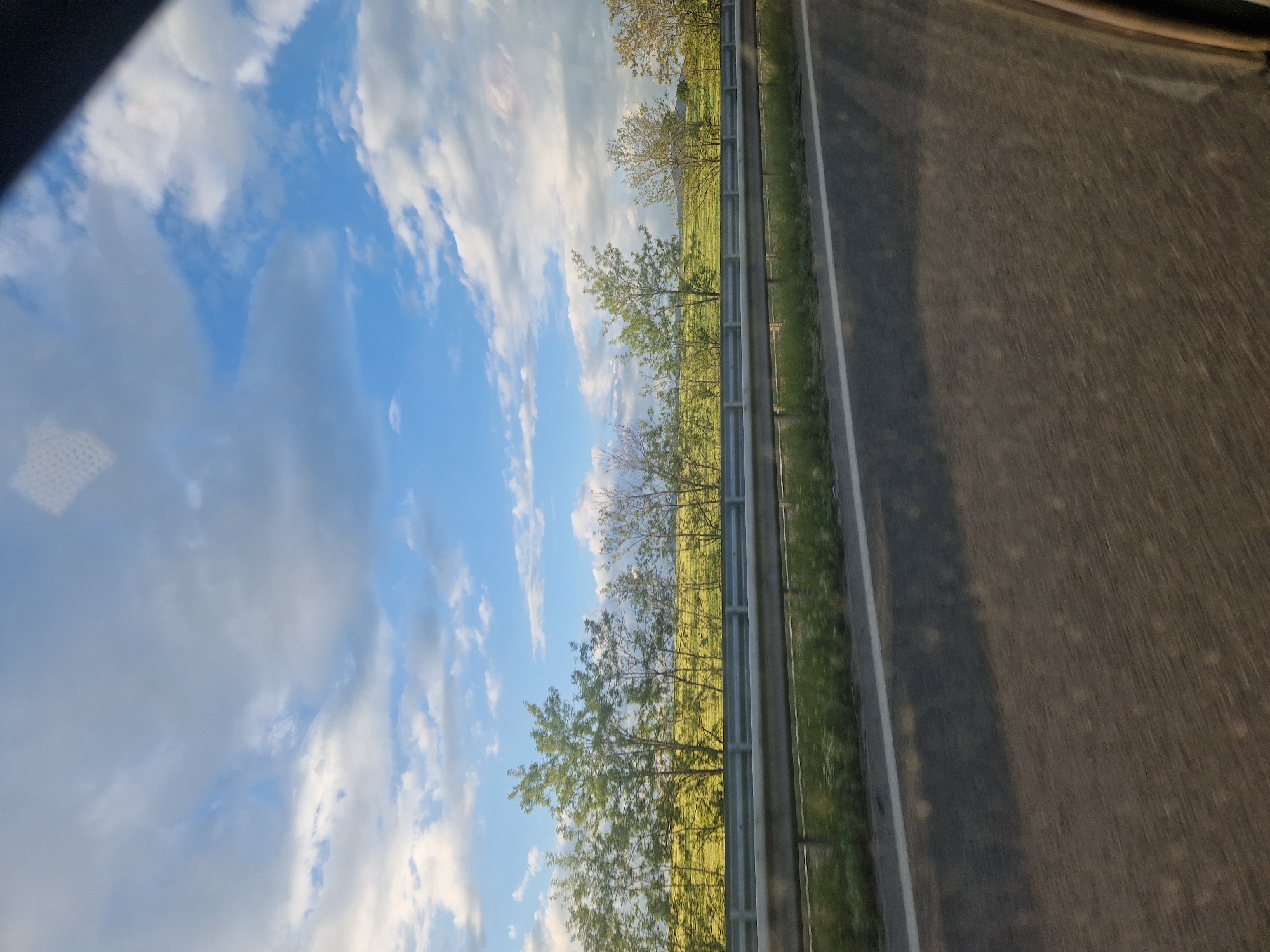 The image depicts a view from inside a vehicle, likely on a highway or major roadway, in an unknown location. A clear sky with scattered clouds fills the upper left portion of the frame, suggesting fair weather. A grassy field with occasional trees runs parallel to the road, separated by a metallic guardrail. The road surface is dark grey, possibly asphalt, and takes up a significant portion of the right side of the image. The shot appears to be taken through a window with some minor distortion or reflection visible. There are no visible people, objects, actions, or interactions that can be discerned in this image other than the scenery and the implied motion of the vehicle.